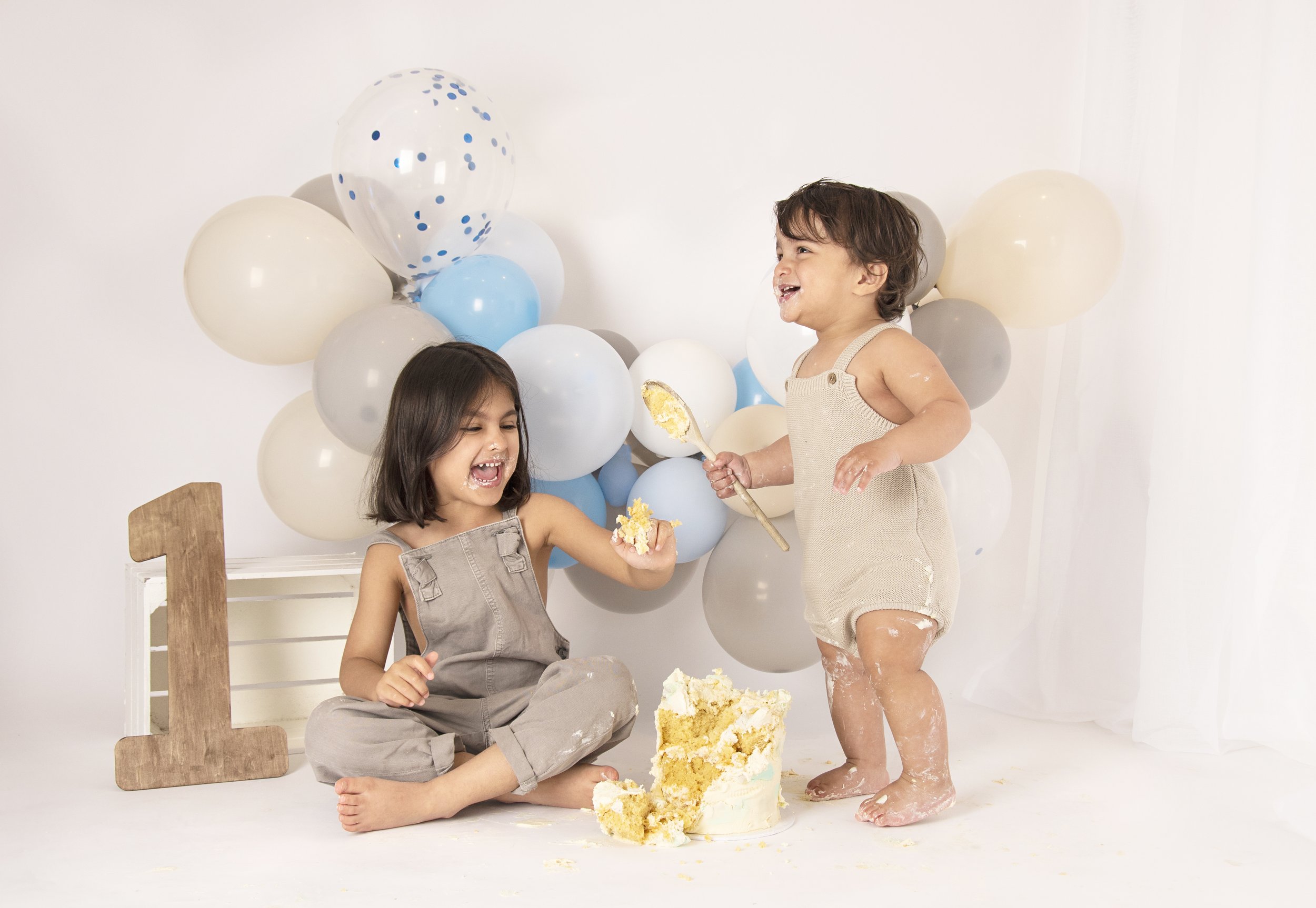 Two children celebrating a first birthday with a cake smash, surrounded by balloons and a wooden number 1.