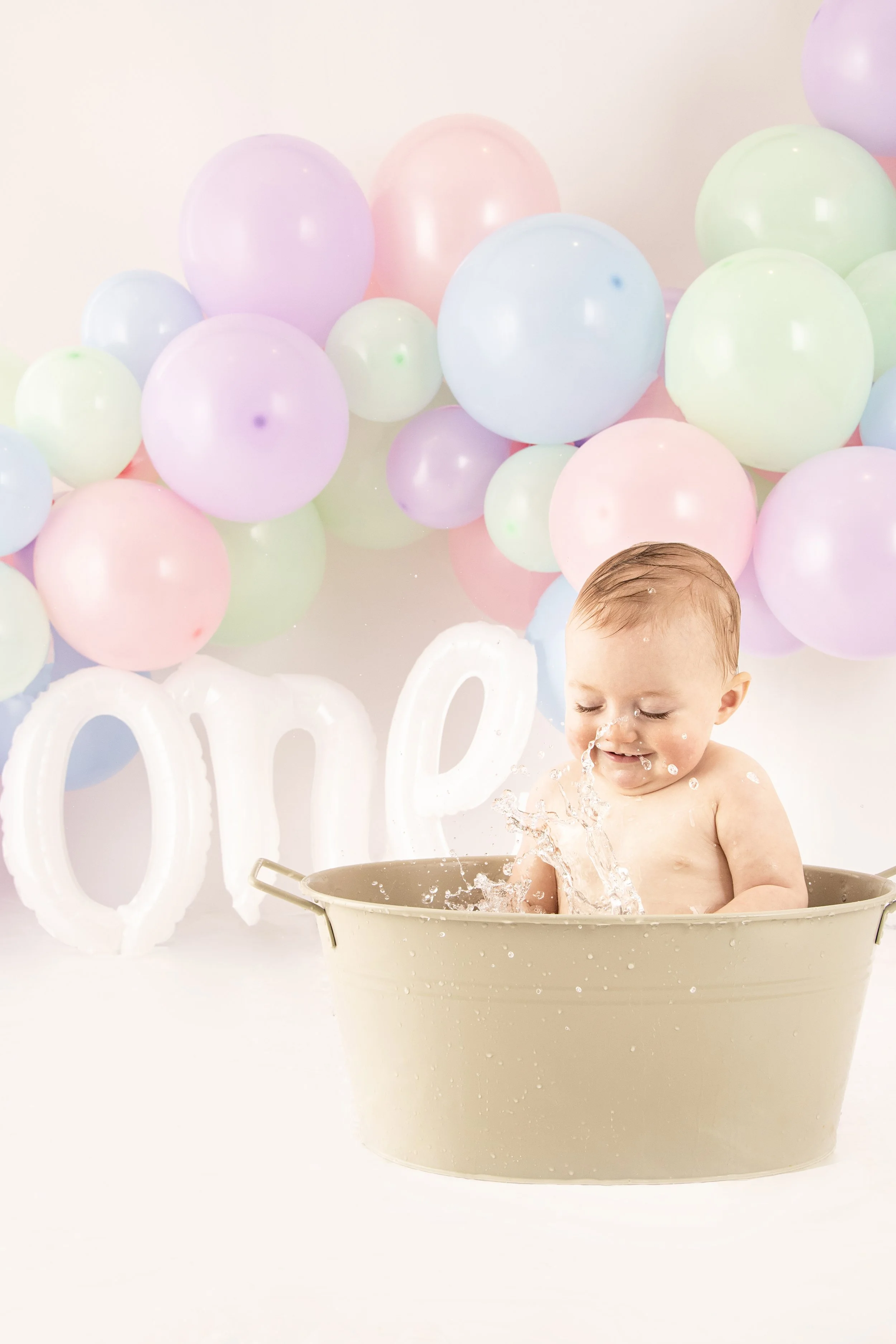 Baby splashing in a bathtub with colorful balloons and the word "one" in the background.