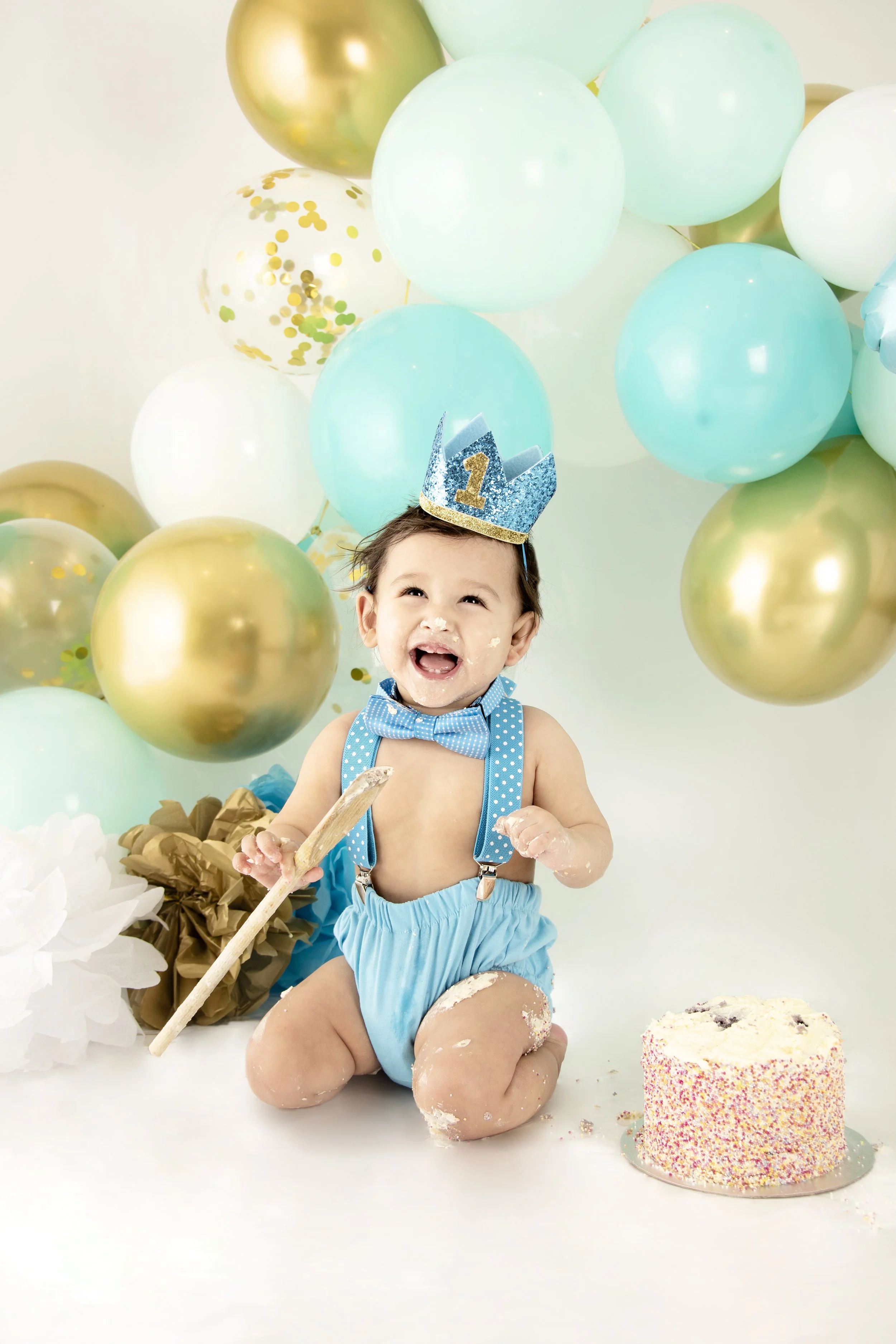 Smiling baby boy with a blue first birthday crown and bow tie, surrounded by blue and gold balloons, sitting next to a cake.
