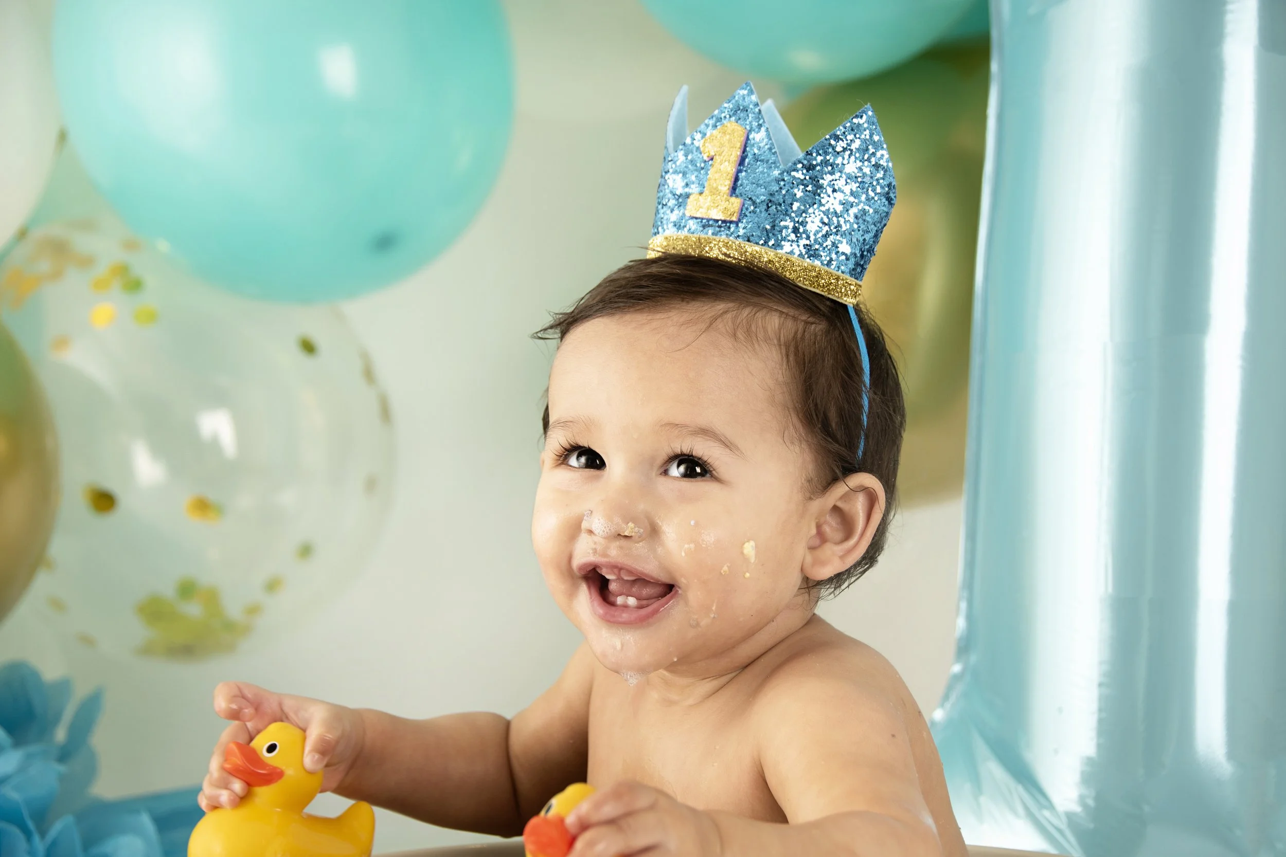 A smiling baby wearing a blue party hat with a number "1" on it, surrounded by colorful balloons, and holding a rubber duck.