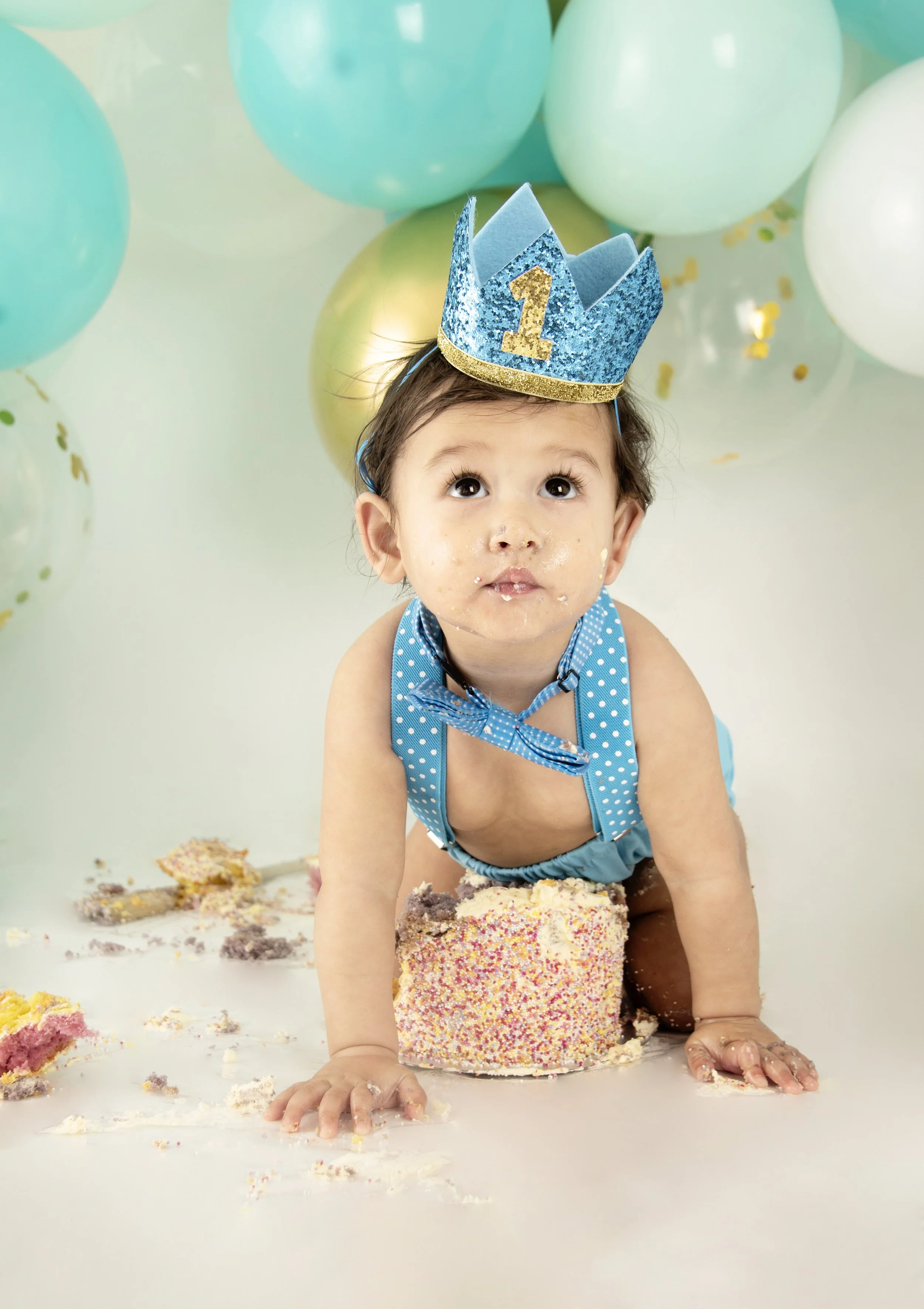 Baby wearing a blue crown and outfit, with cake and balloons, celebrating first birthday.