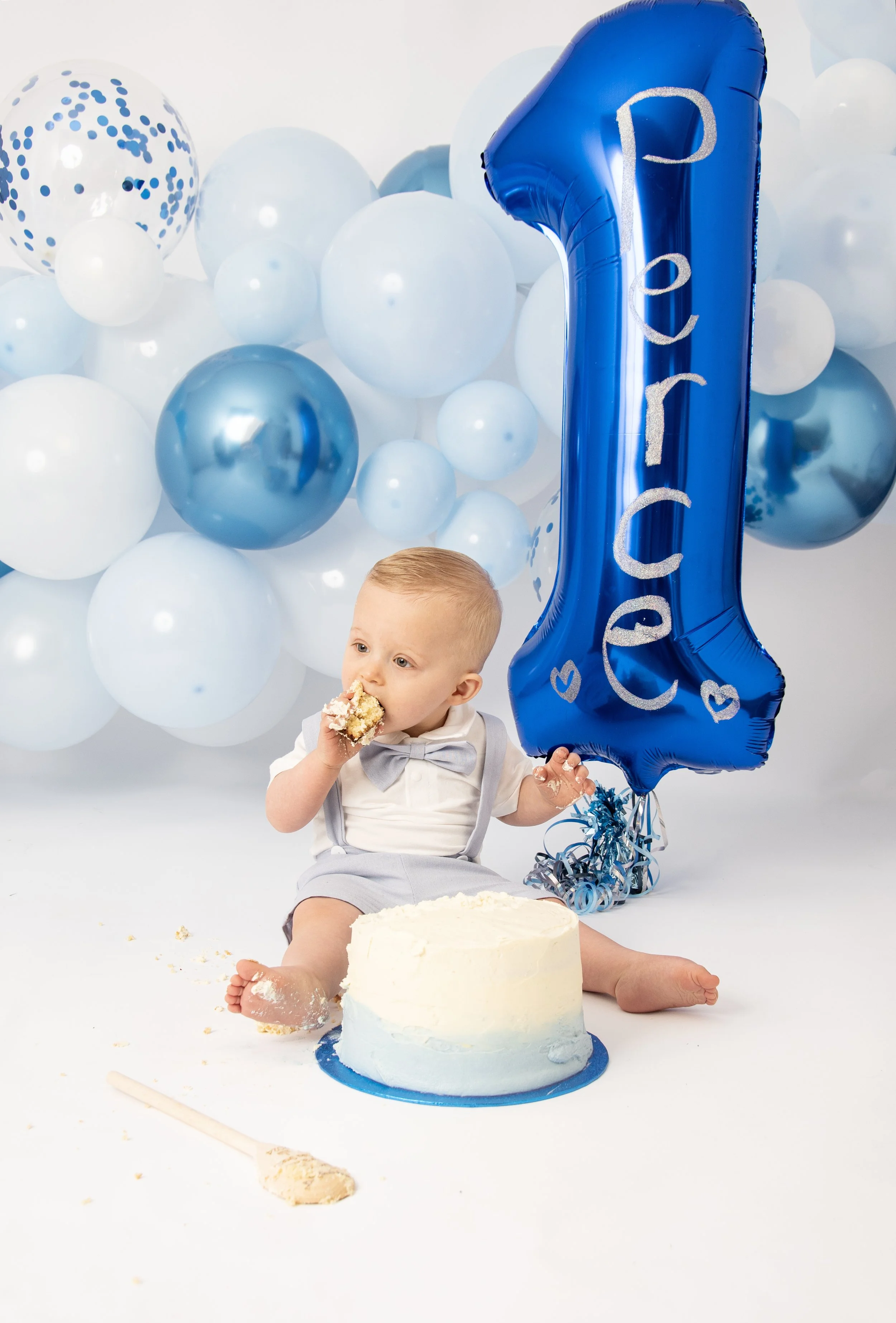 Baby in light blue outfit eating cake next to a blue "1" balloon, surrounded by blue and white balloons.