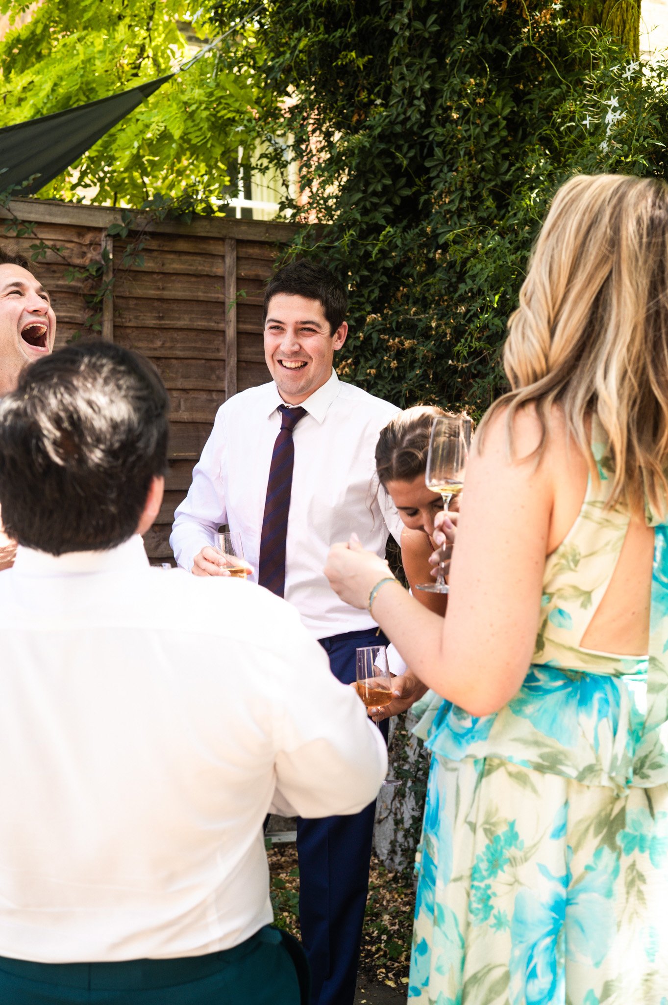 Group of people celebrating outdoors, holding wine glasses, smiling, and engaged in conversation, with greenery and a wooden fence in the background.