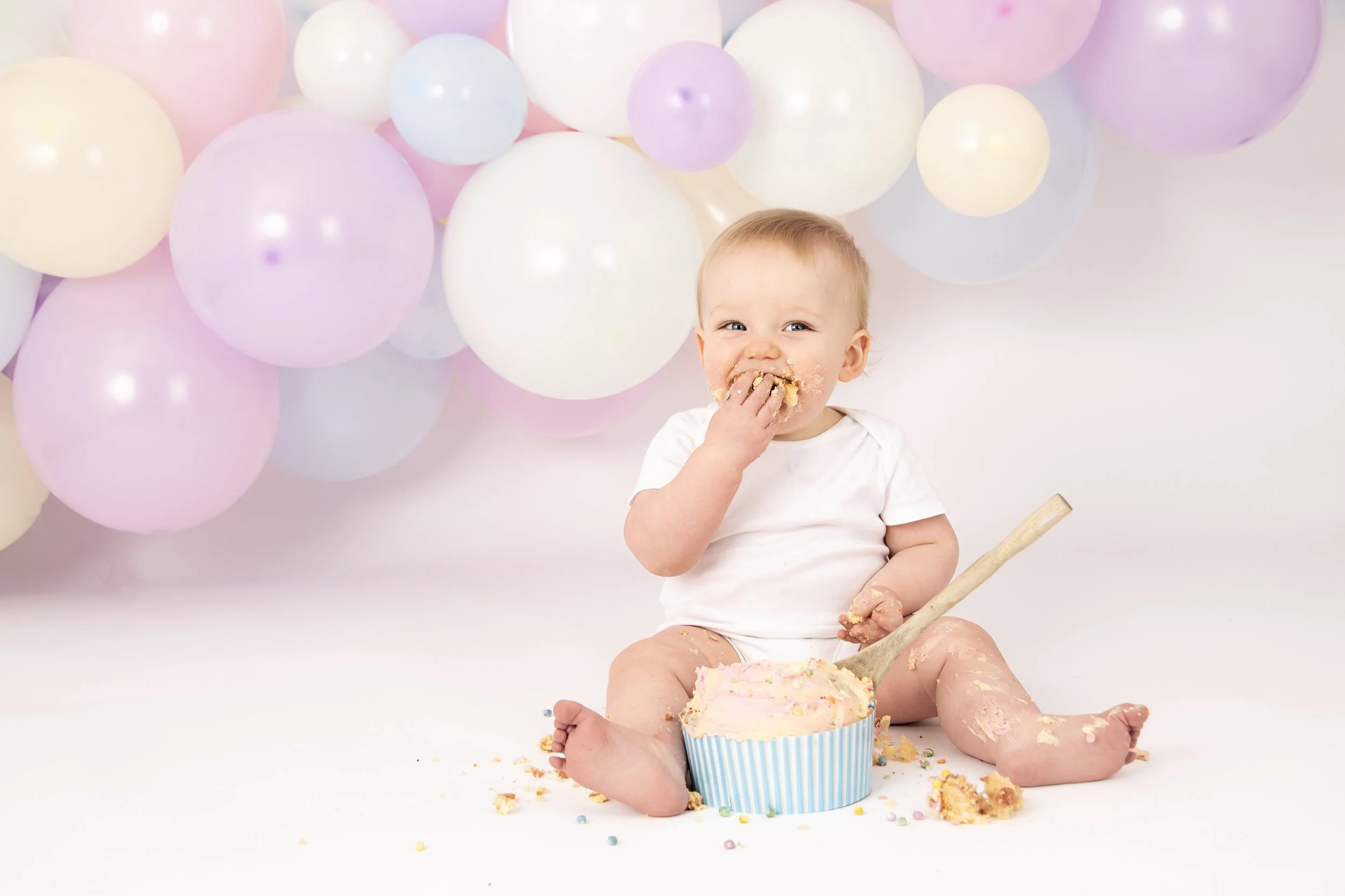 Baby enjoying a cake during a cake smash session with pastel balloons in the background.