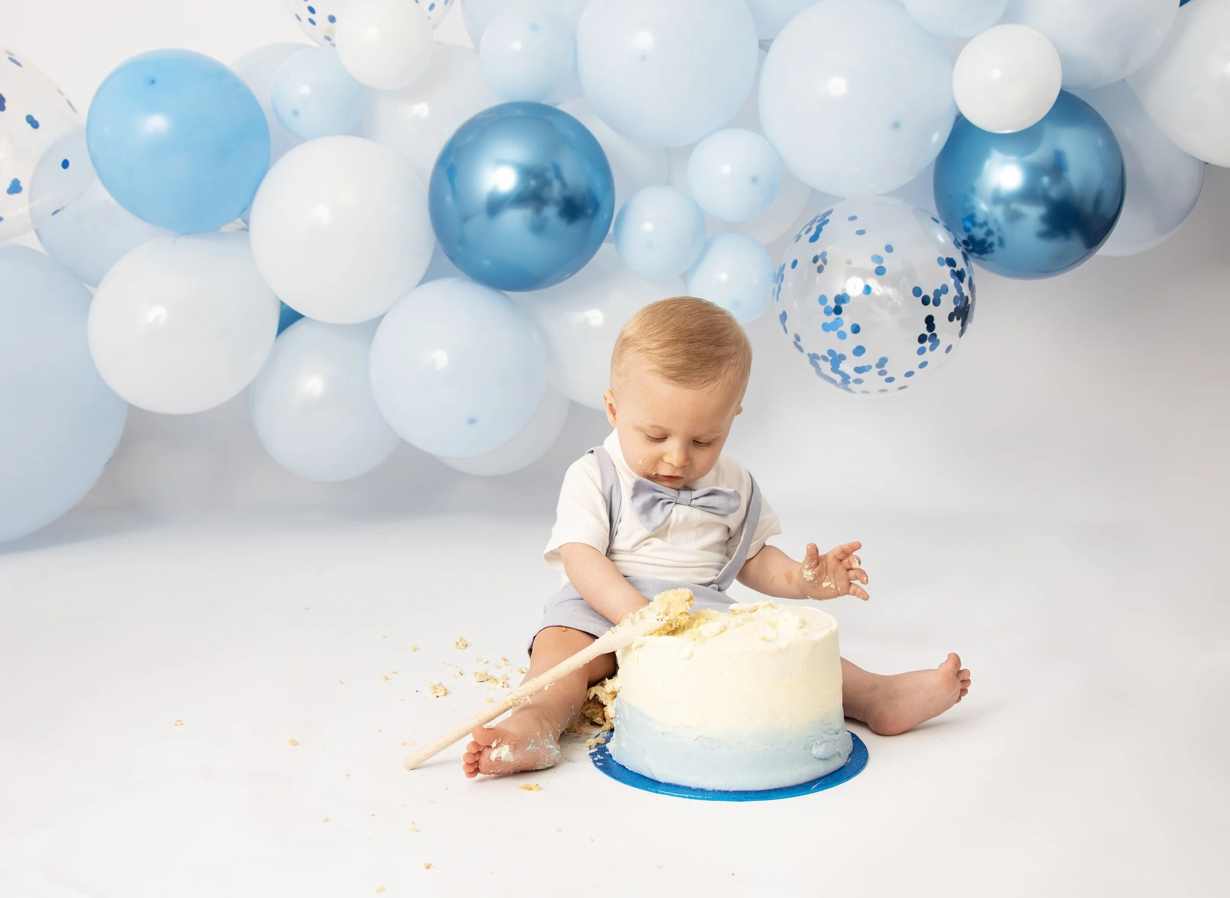 Baby sitting among blue and white balloons with a partially smashed cake.