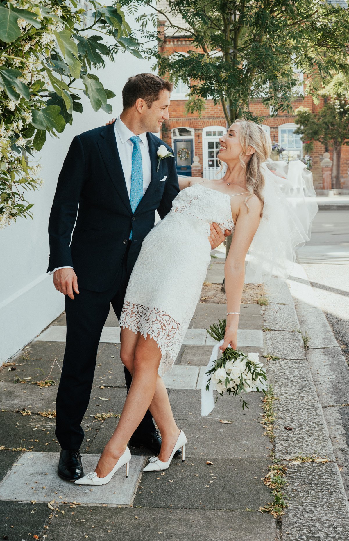 A groom holding a bride who is holding a bouquet of white flowers, in an outdoor setting with trees and buildings in the background.