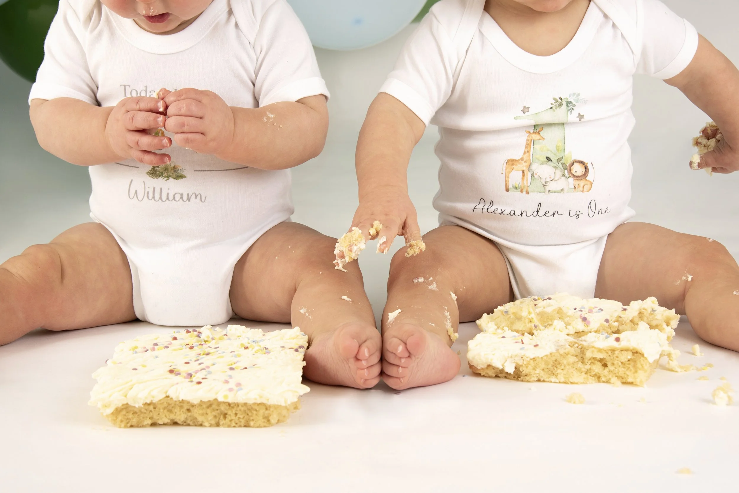 Two babies in white onesies sitting on the floor with smashed cakes, having a cake smash photoshoot.
