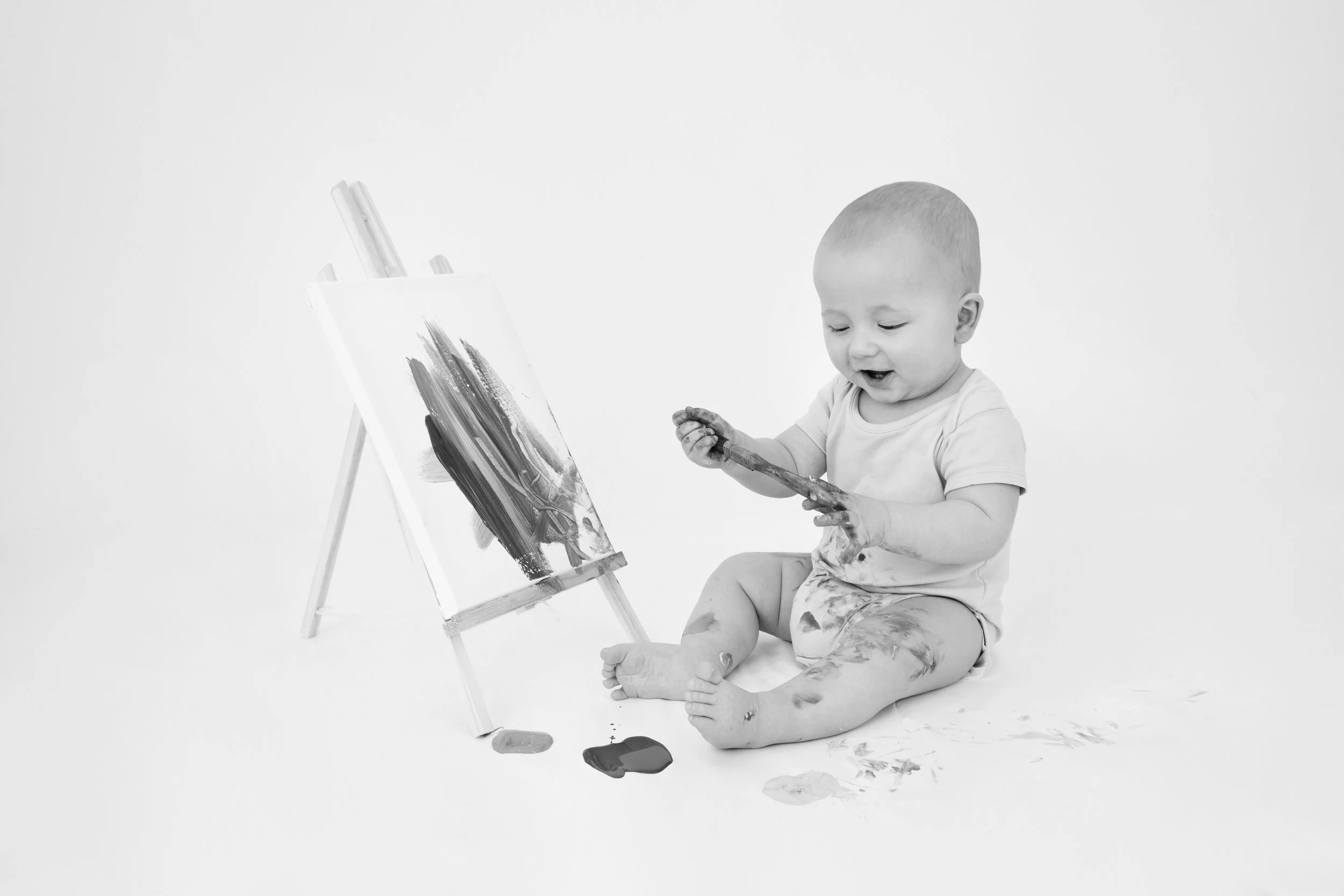 Baby sitting on floor with paint-covered hands and feet painting on canvas on easel.