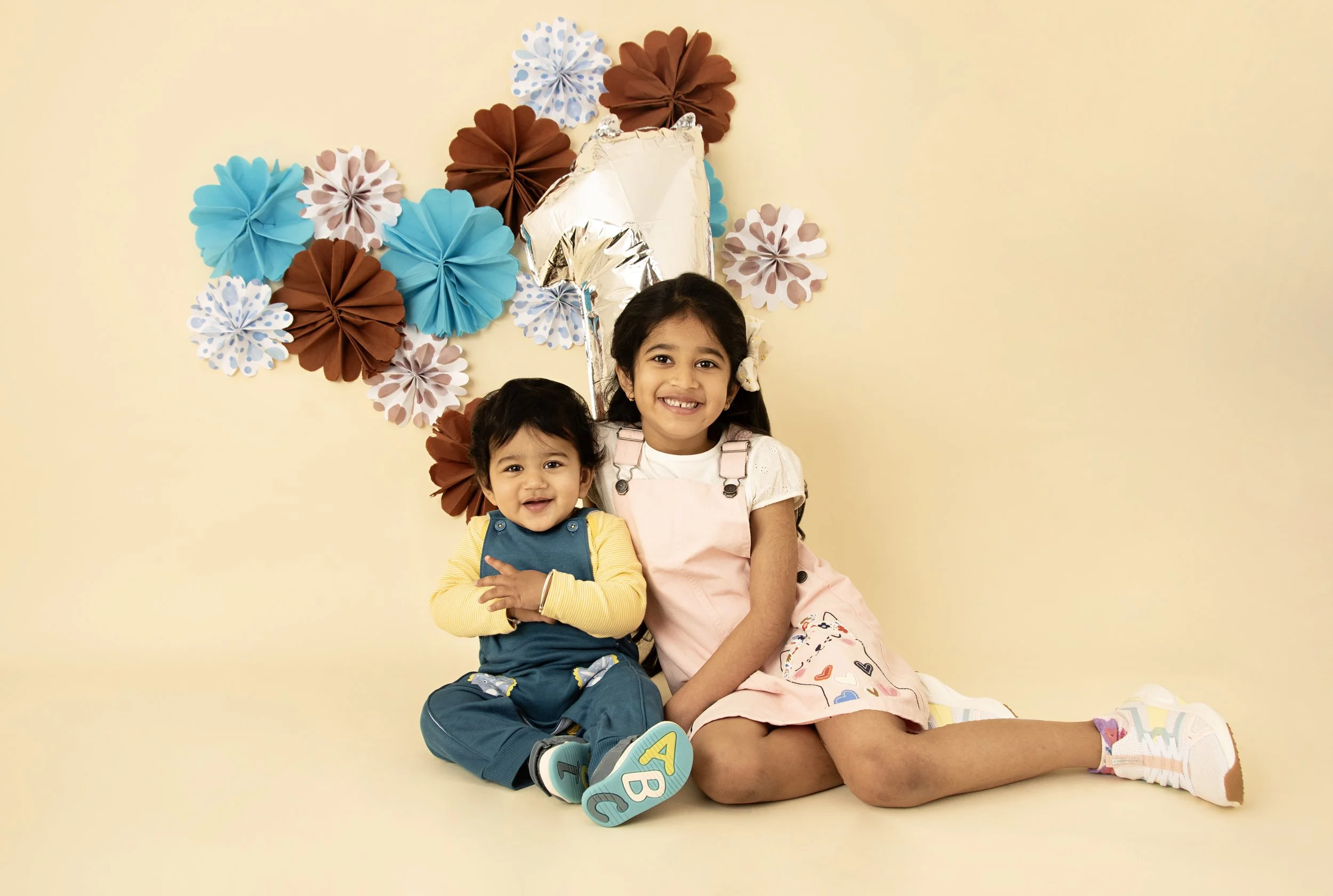 Two children sitting in front of a decorative background with paper flowers and a silver balloon shaped like the number one. The older child is wearing a pink dress, and the younger child is in a blue outfit, both smiling.