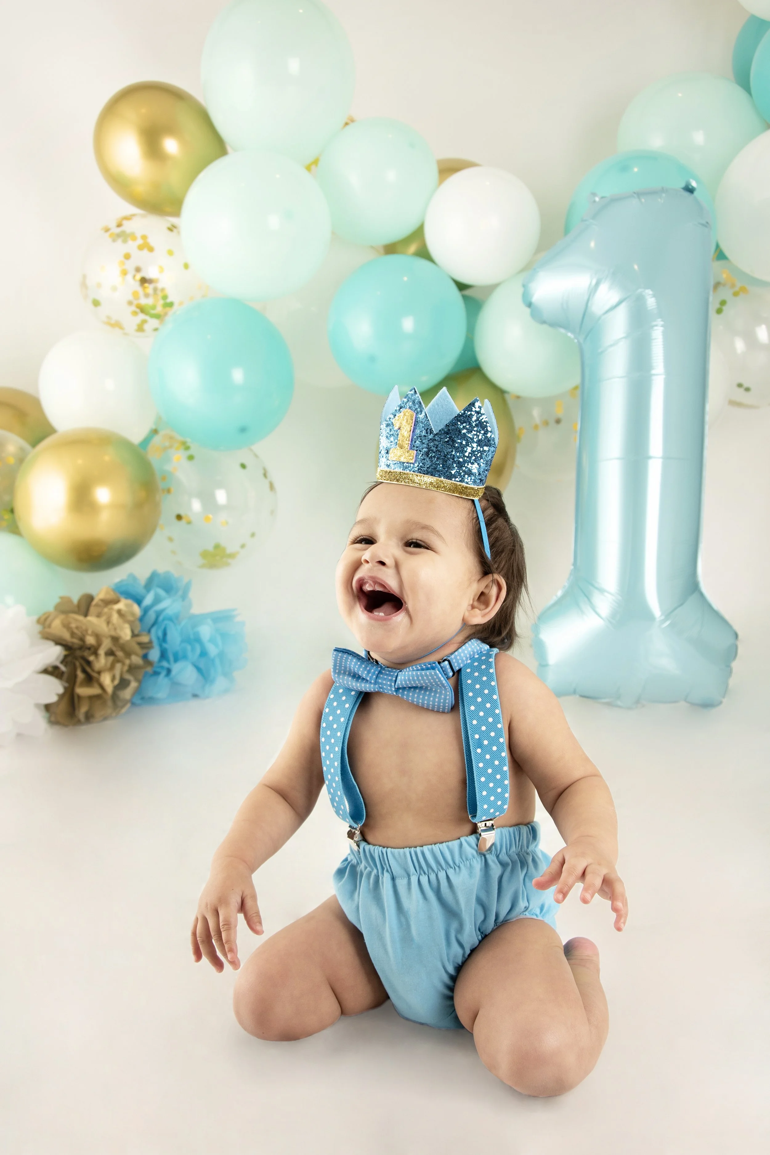 A baby in blue suspenders and a party hat celebrates their first birthday with balloons and a large number one balloon.