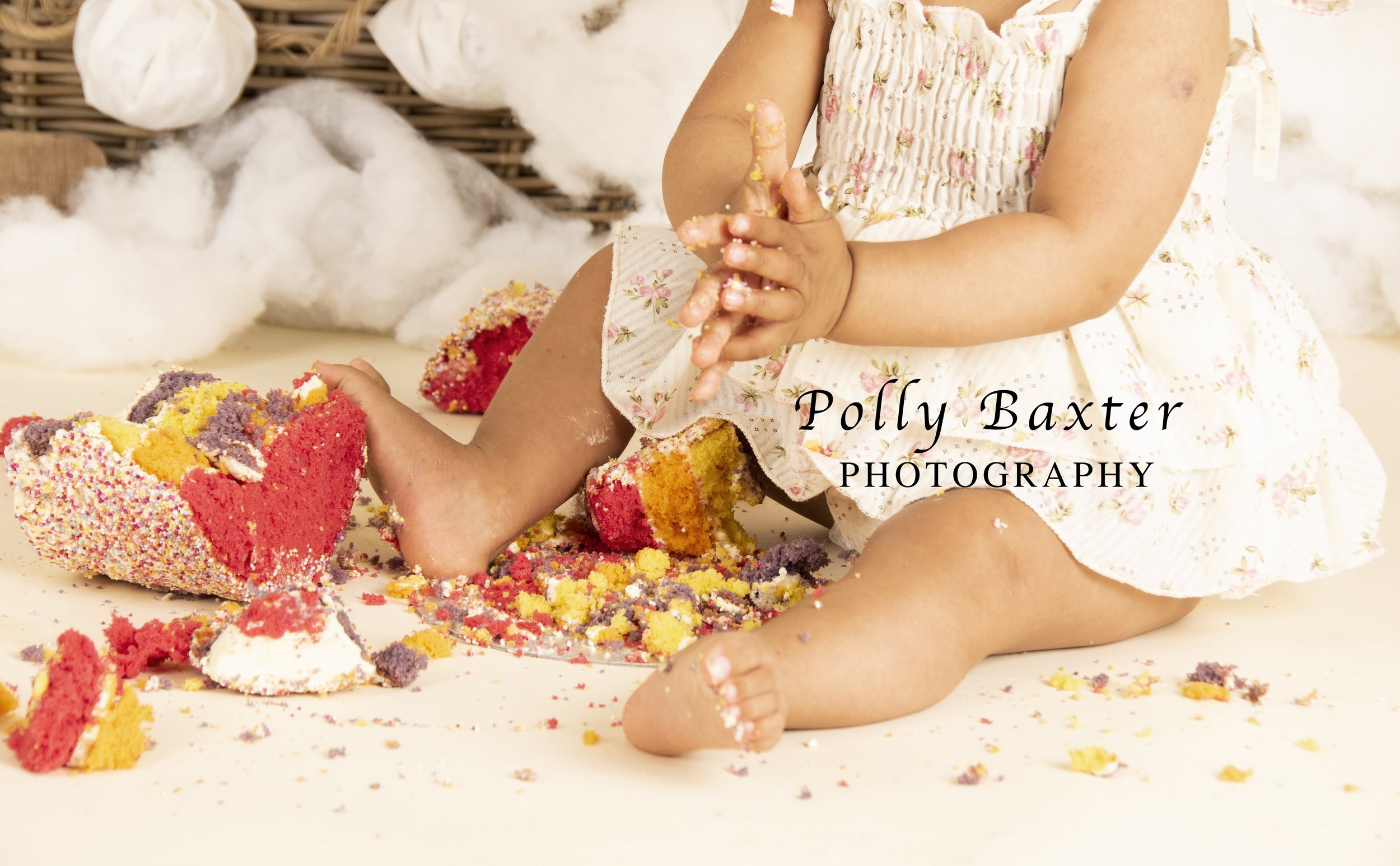 A baby in a floral dress sitting on the floor playing with a smashed colorful cake, surrounded by fluffy decorations.