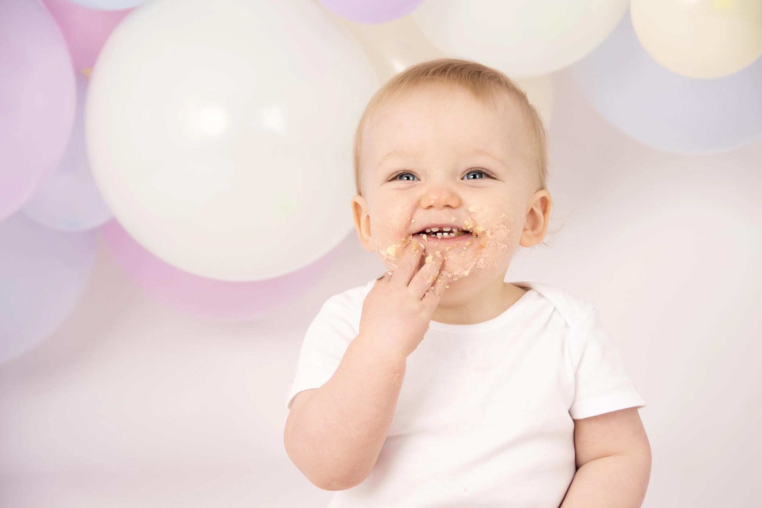 Smiling baby with cake on face and hand, surrounded by pastel balloons.