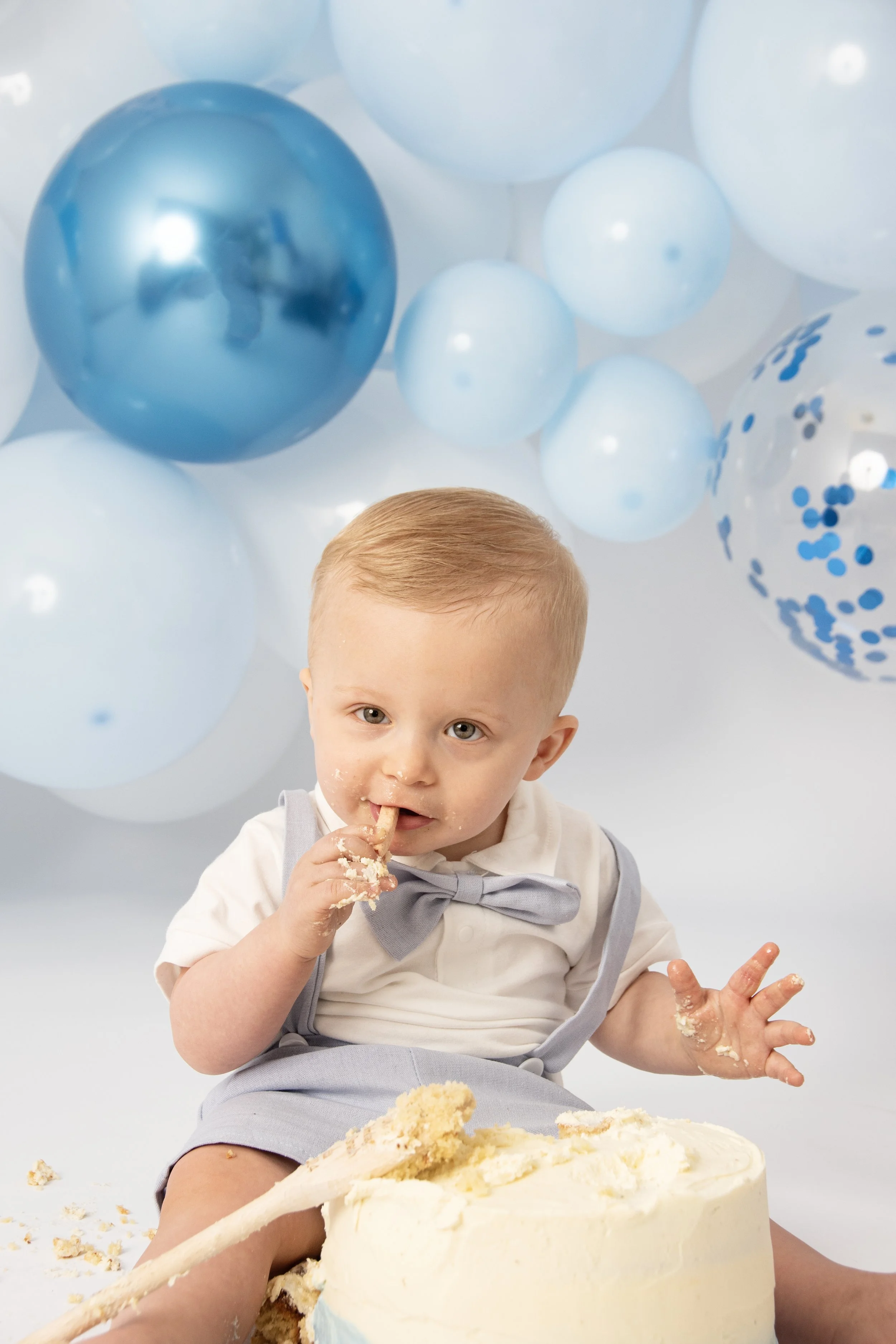 A baby in a white shirt and blue bow tie is sitting on the floor with a cake covered in white frosting in front of him. The baby is eating the cake with his hands, and there are blue and white balloons in the background.
