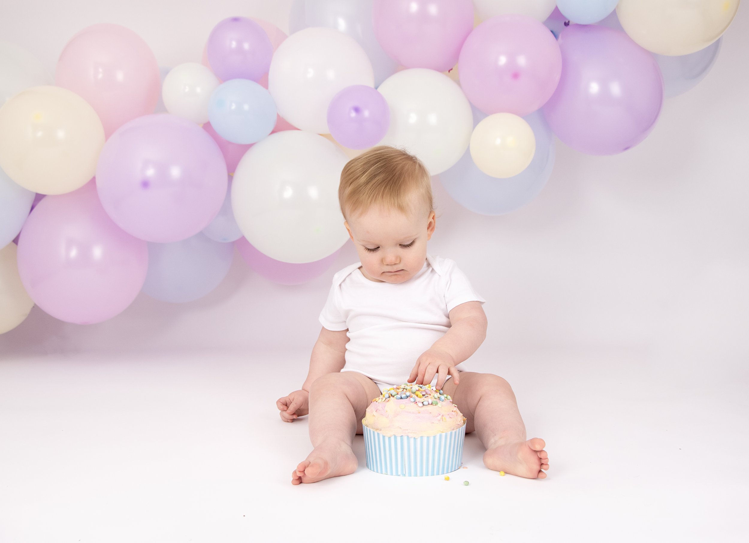 Baby sitting in front of pastel balloons and examining a large cupcake with colorful sprinkles.