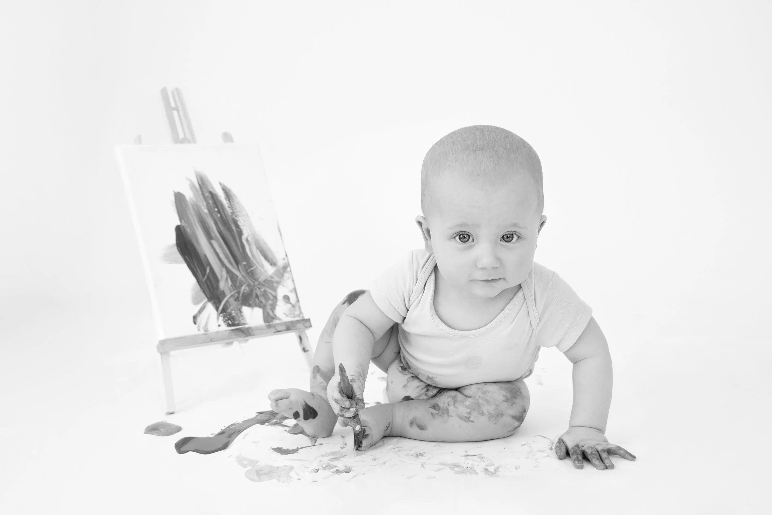 Black and white image of a baby sitting on the floor with paint on their hands and legs, next to an easel with a painted canvas.