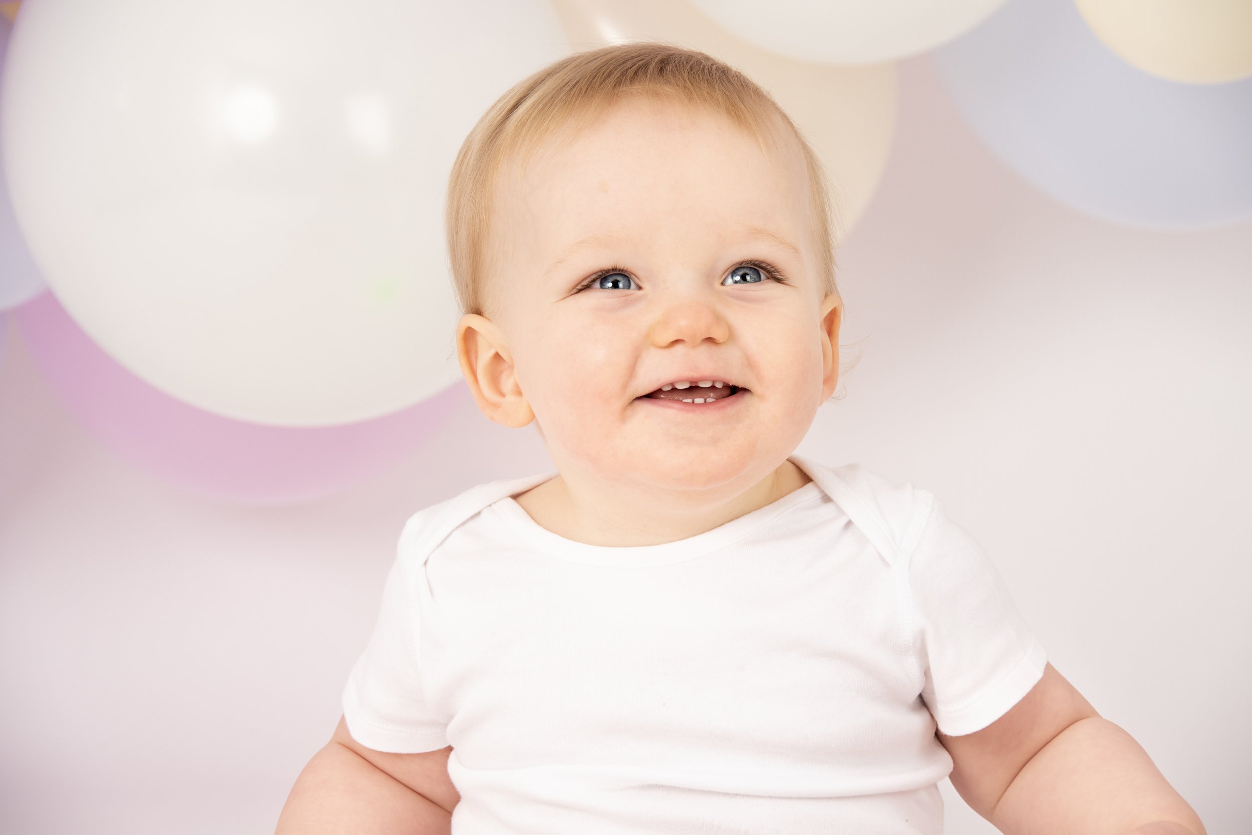 Smiling baby in a white shirt with pastel balloons in the background.
