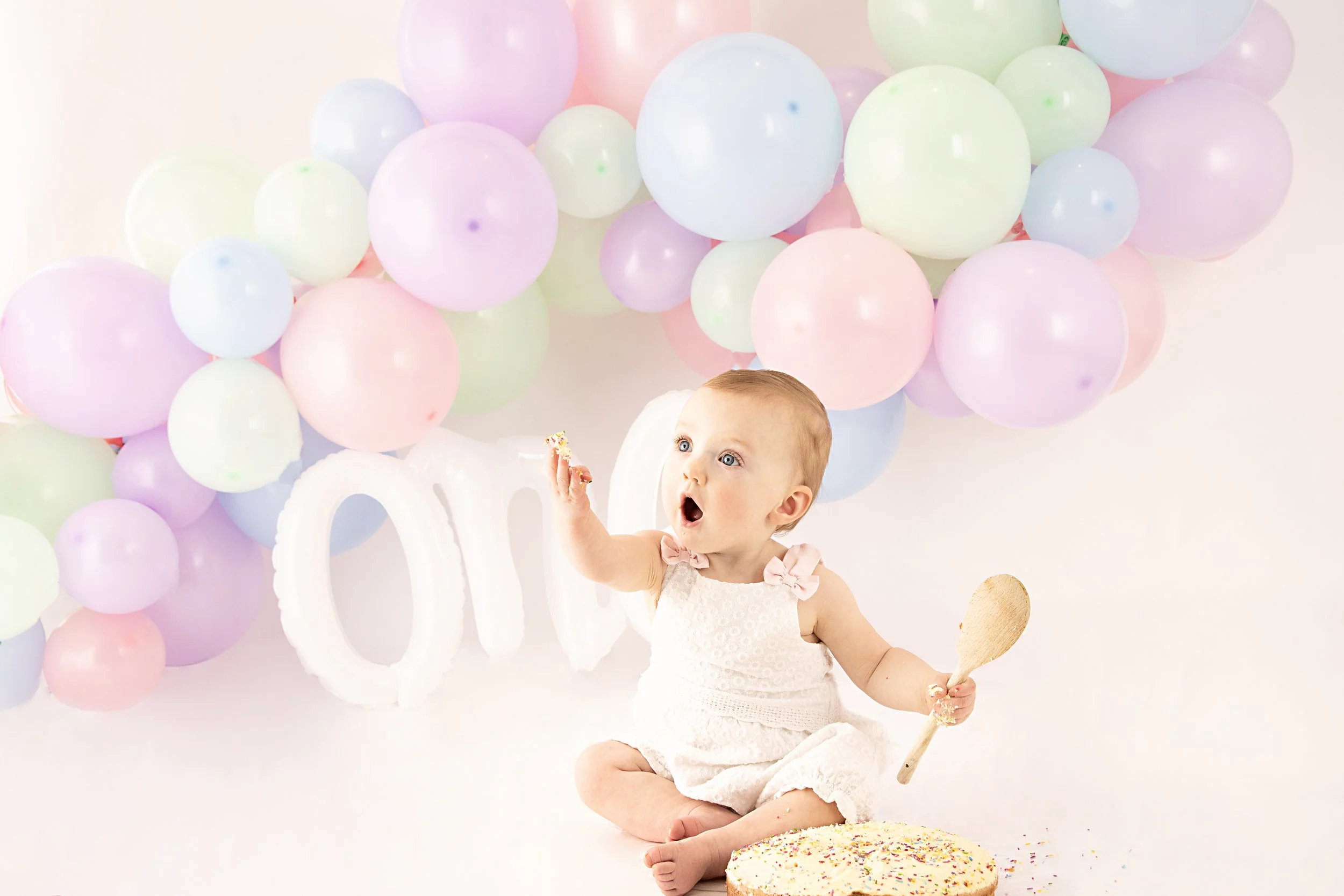 Baby in white outfit sitting with cake and pastel balloons, holding a wooden spoon, against a "one" backdrop.