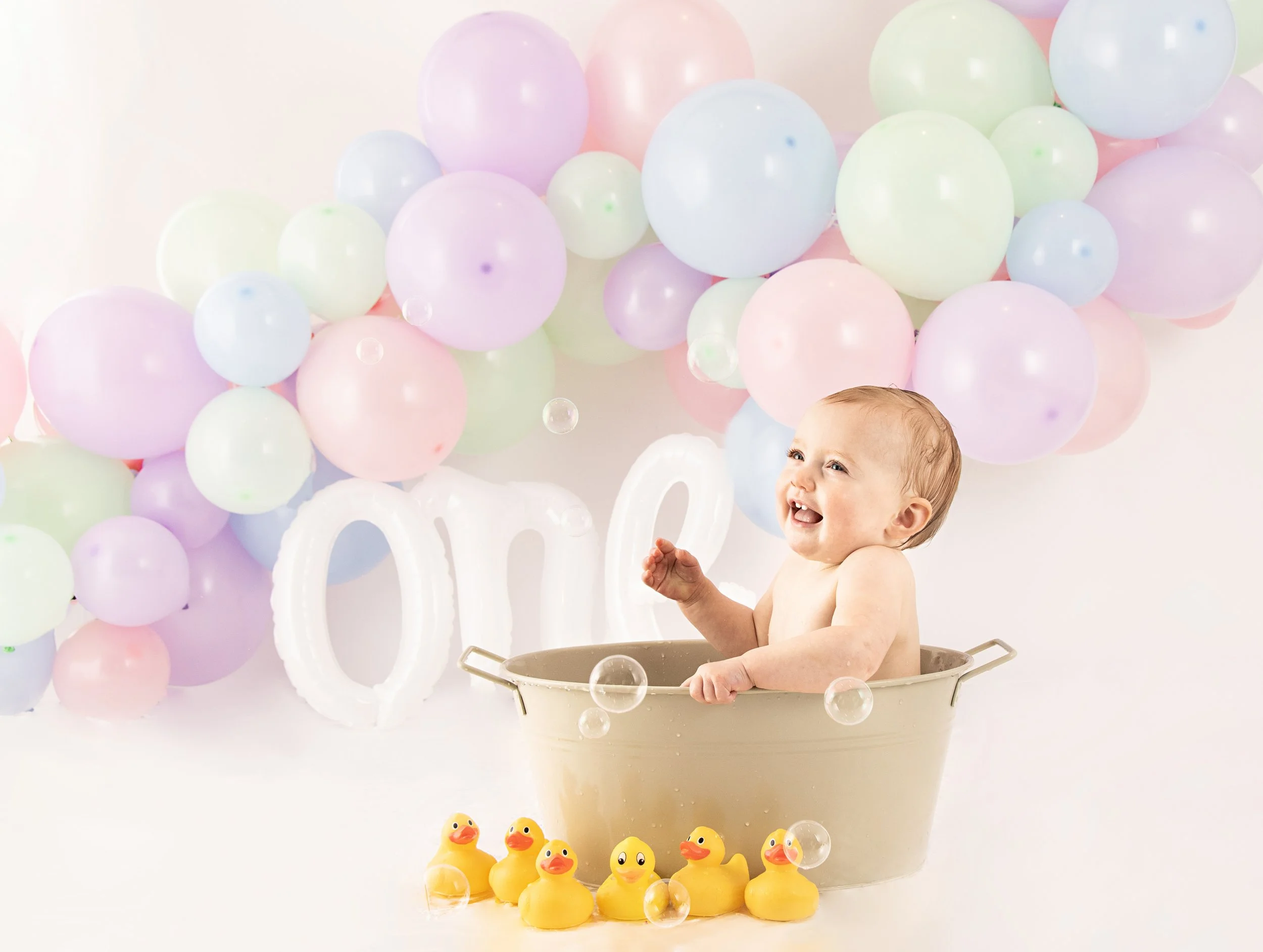 Smiling baby in a bathtub with rubber ducks, colorful pastel balloons, and "One" sign in background.