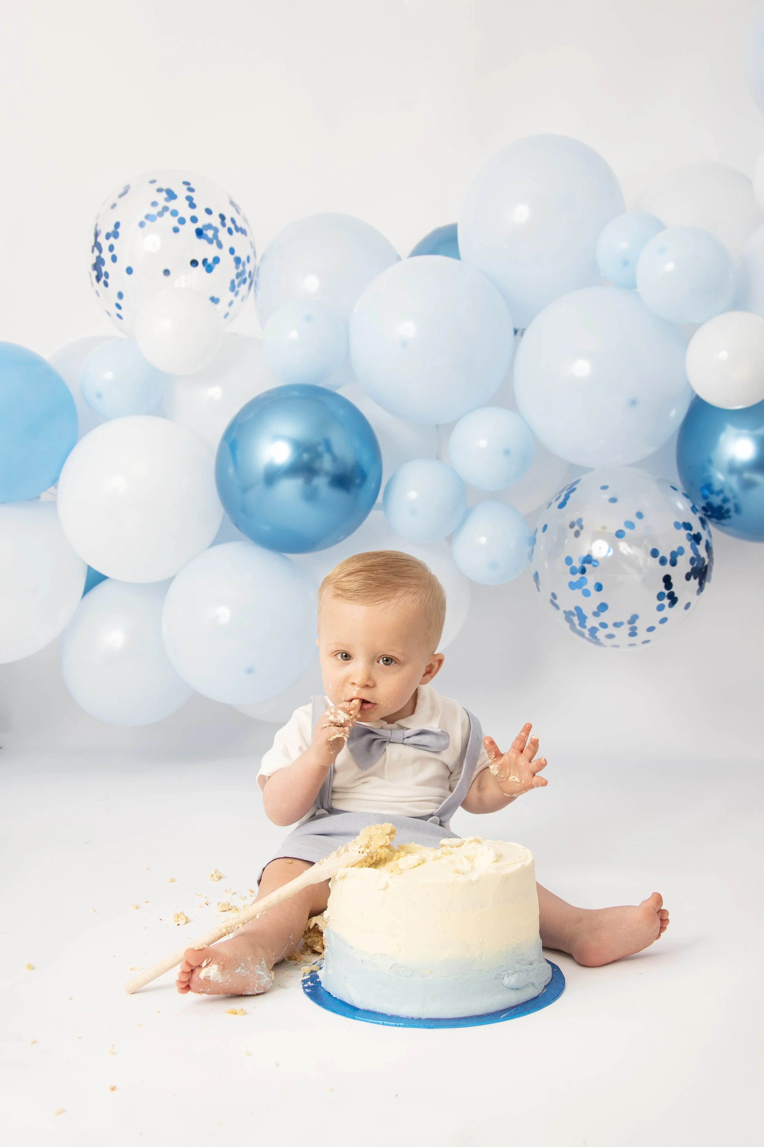 Baby sitting on floor with a cake, surrounded by blue and white balloons, eating with hand.