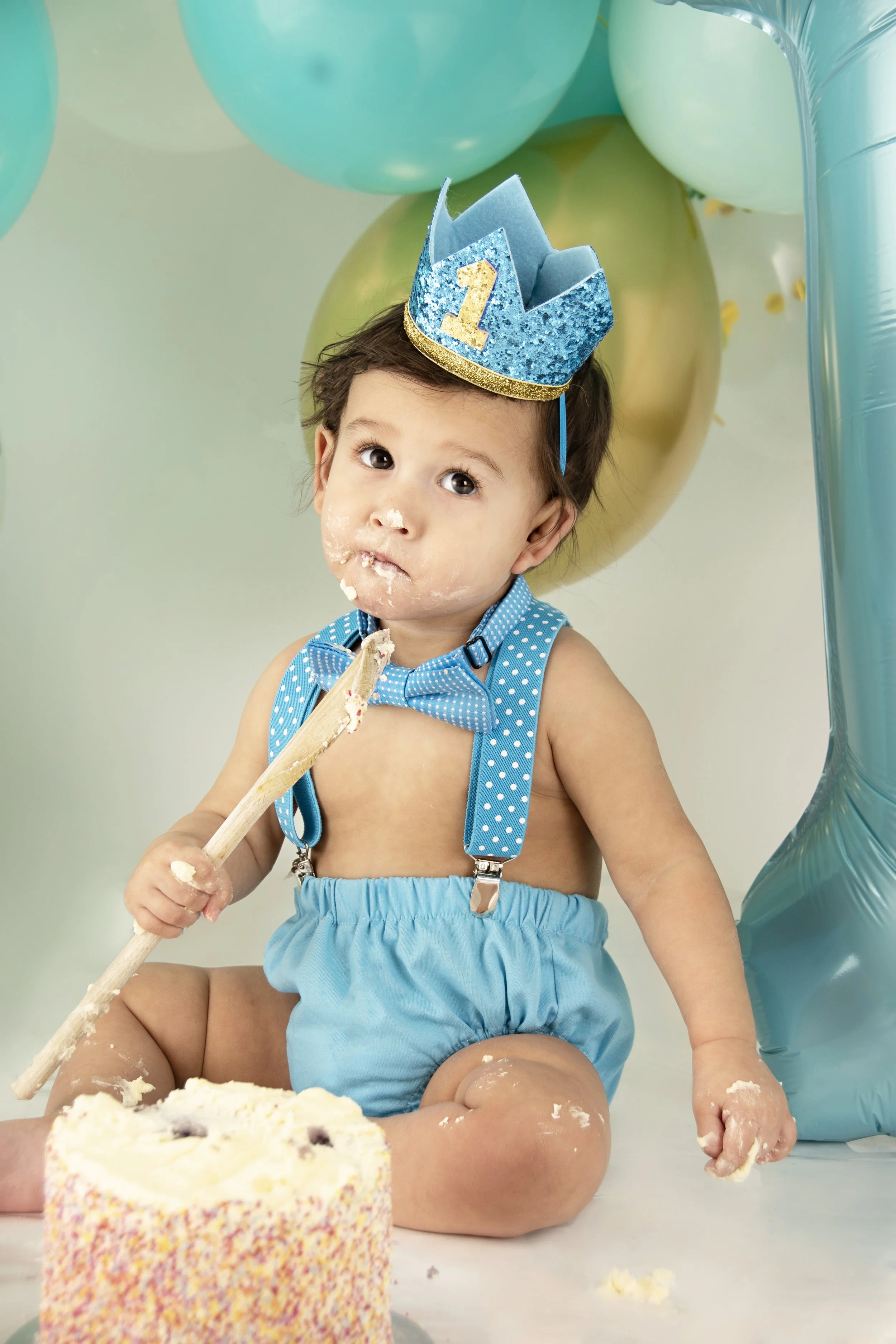 Baby celebrating first birthday in blue outfit with party hat and cake. "1" balloon and colorful balloons in background.