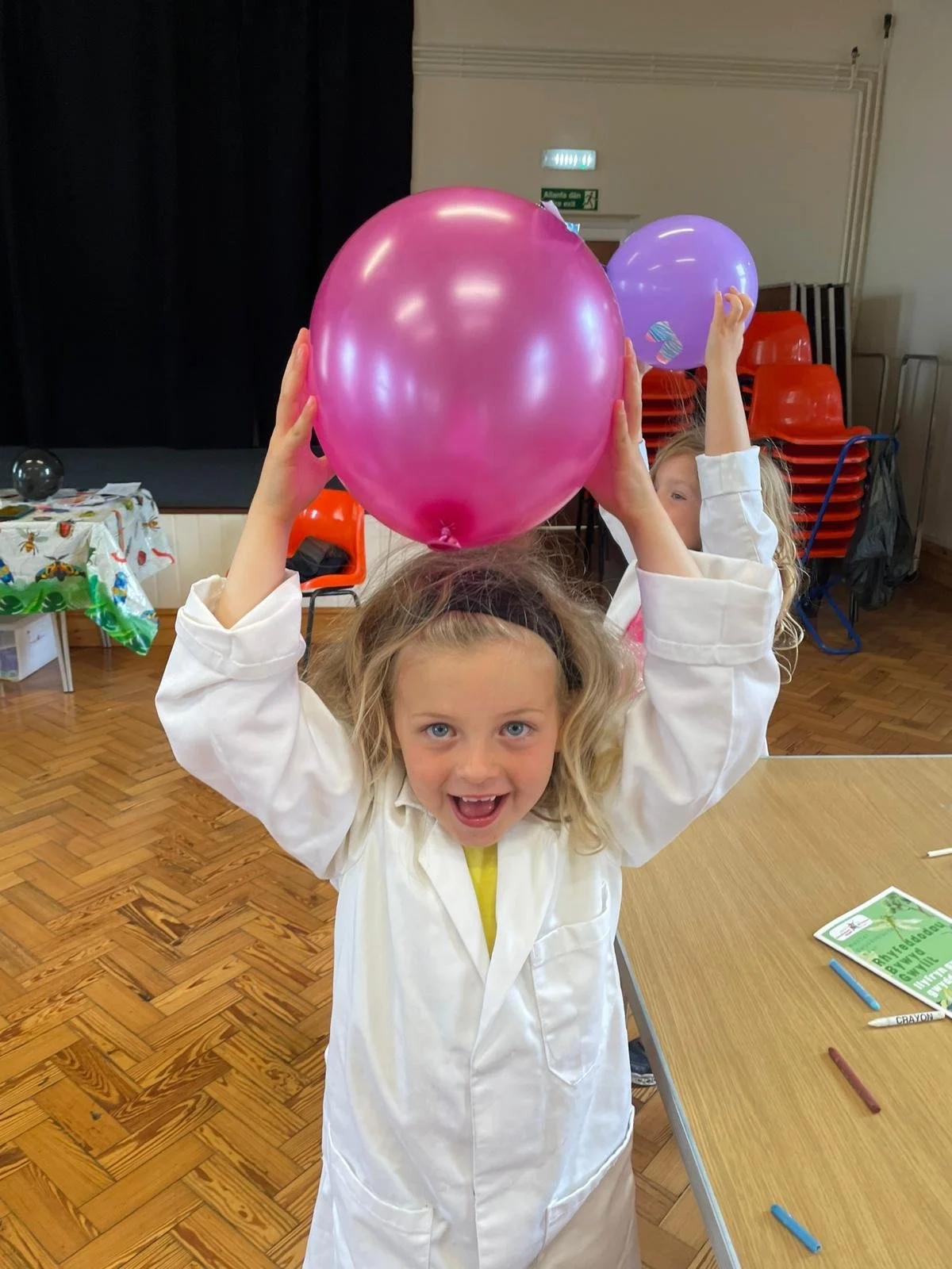 A young girl in a white lab coat holding a pink balloon above her head with a big smile in a classroom or event hall, with other children and balloons in the background.