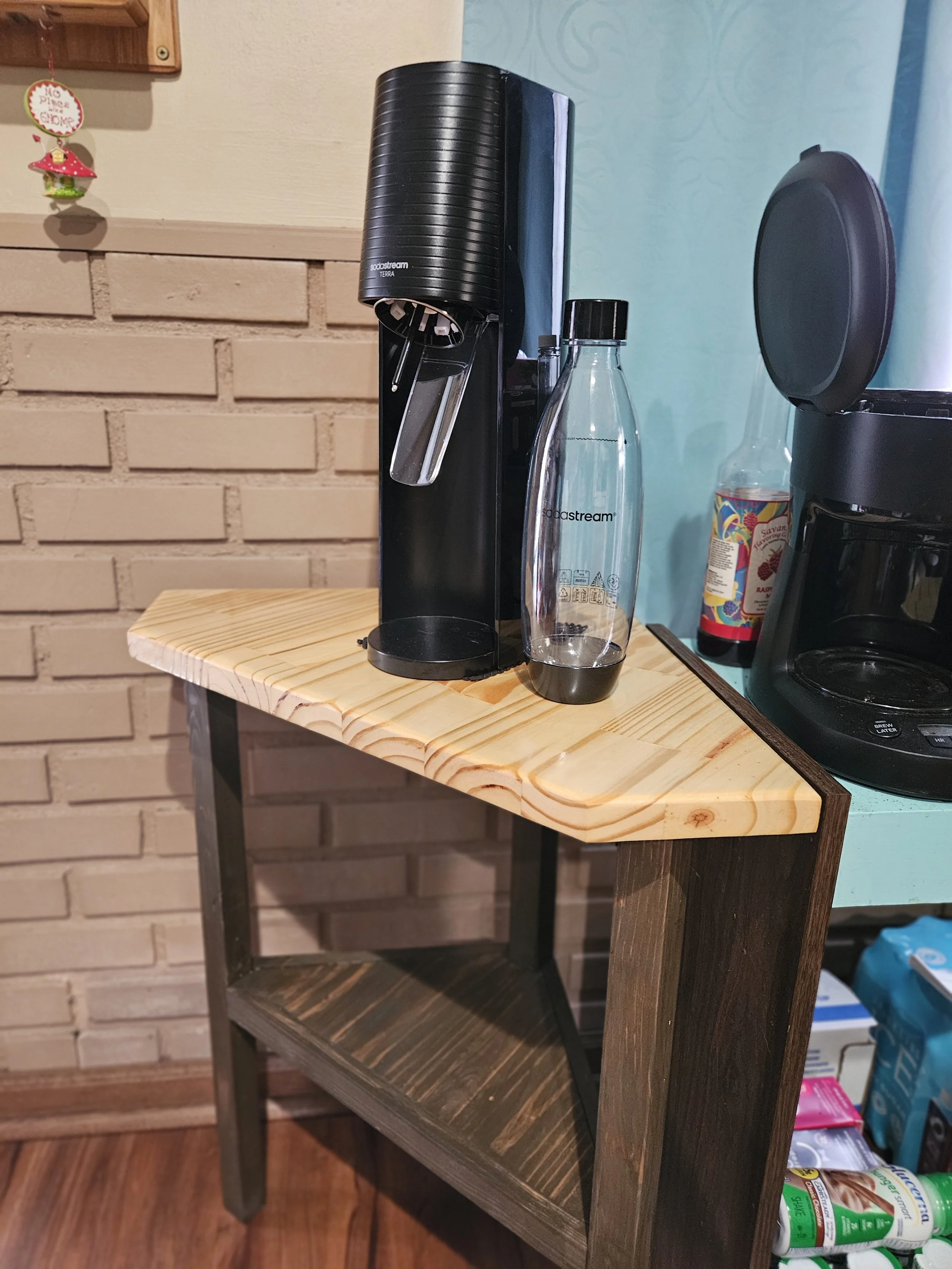 A wooden table with a black sodaStream home soda maker, a bottle of sodaStream water, and a black coffee maker, against a brick wall and blue wall background.