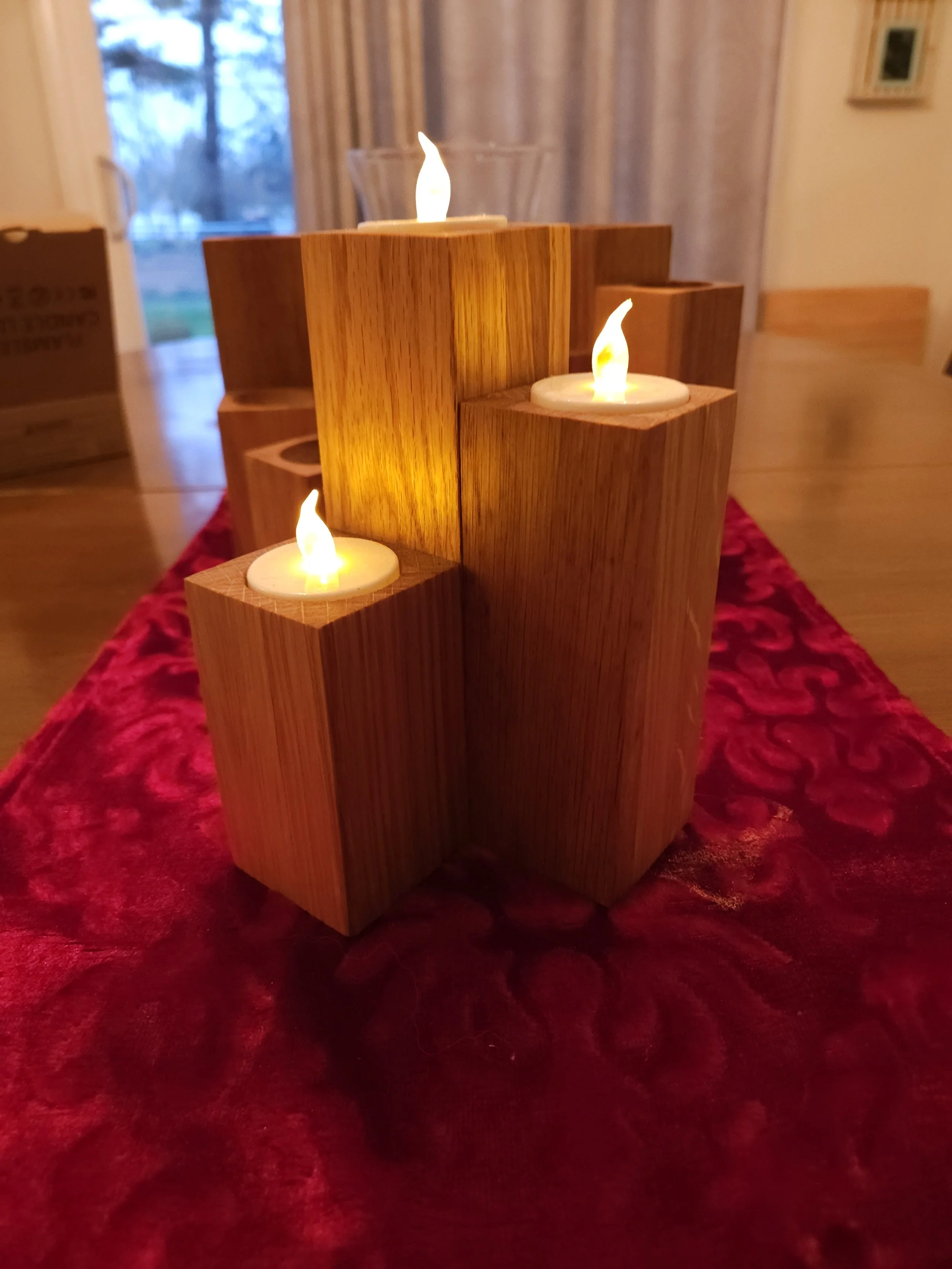 Wooden candle holder with four electric candles on a red velvet table runner inside a room with curtains and a view outside window.