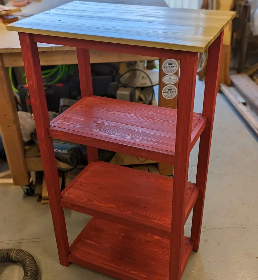 A four-tier wooden shelf with the top shelf painted blue and the lower shelves painted red, standing in a workshop.