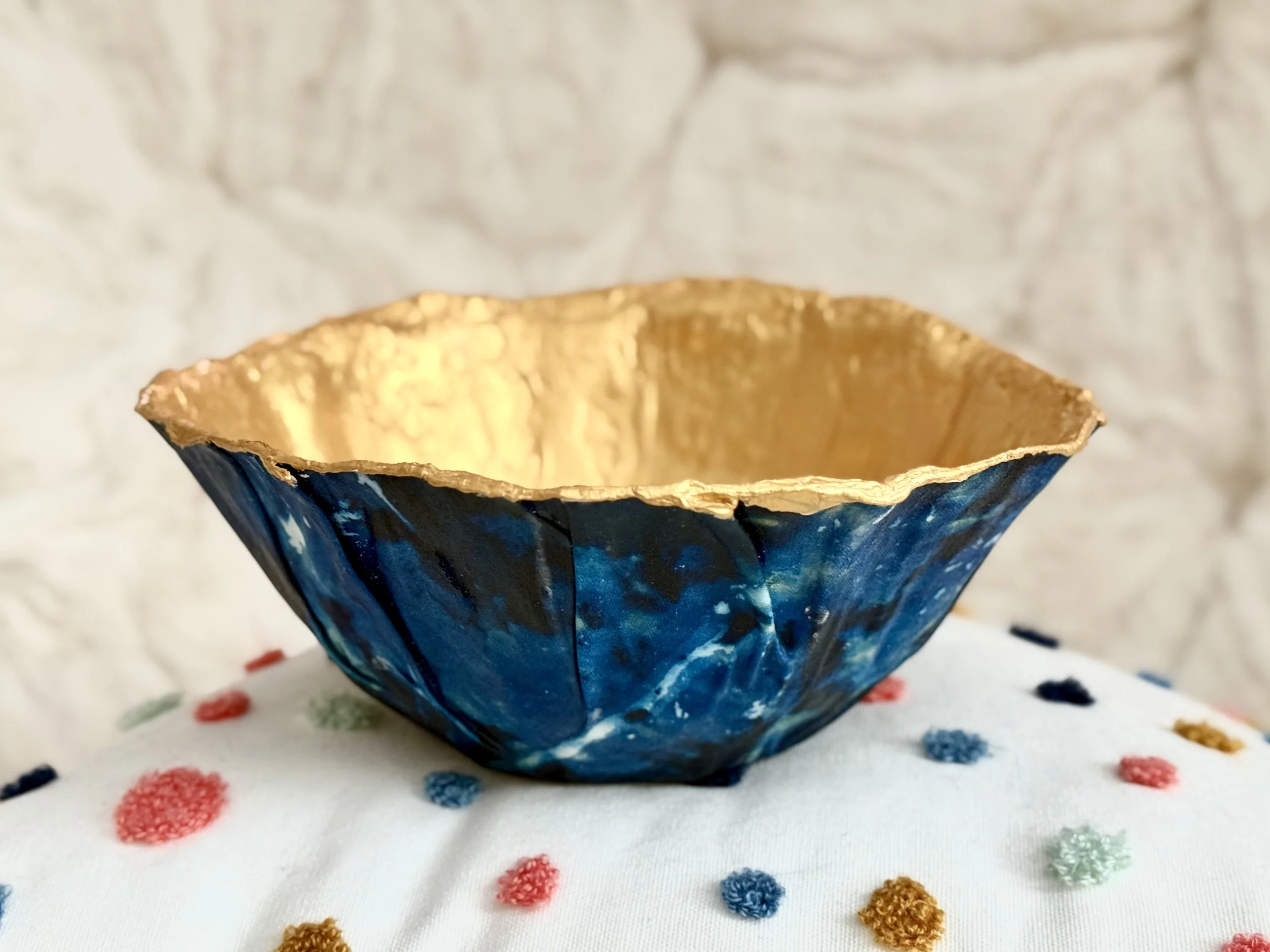 Close-up of a handmade bowl with a gold interior and a blue marbled cyanotype fabric exterior, placed on a white fabric with colorful embroidered polka dots, against a light-colored stone wall background.