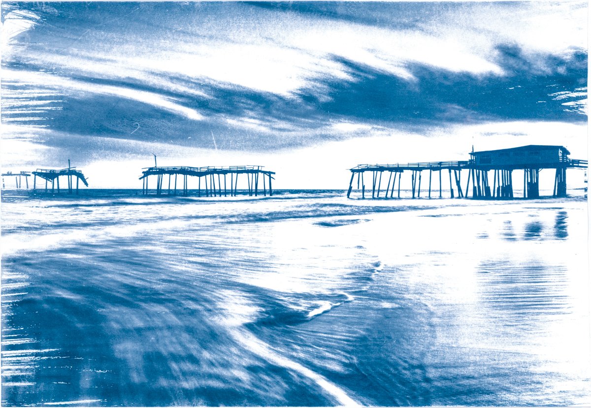 Cyanotype of a silhouetted pier structures extending into the ocean under a partly cloudy sky, with water and waves in the foreground.