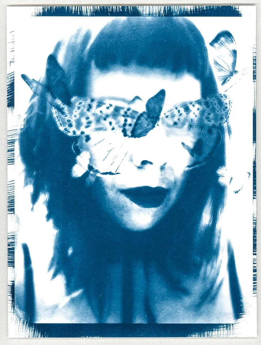 Cyanotype of a woman with shoulder-length hair and dark lipstick has butterflies on her face and around her eyes, with some on her shoulders. The image is in blue and white tones.
