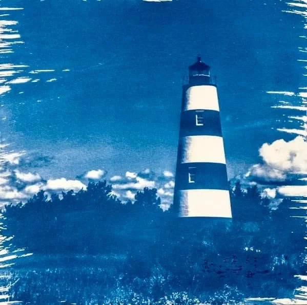 Blue-toned cyanotype photo of a tall striped lighthouse on Sapelo Island surrounded by clouds and water.