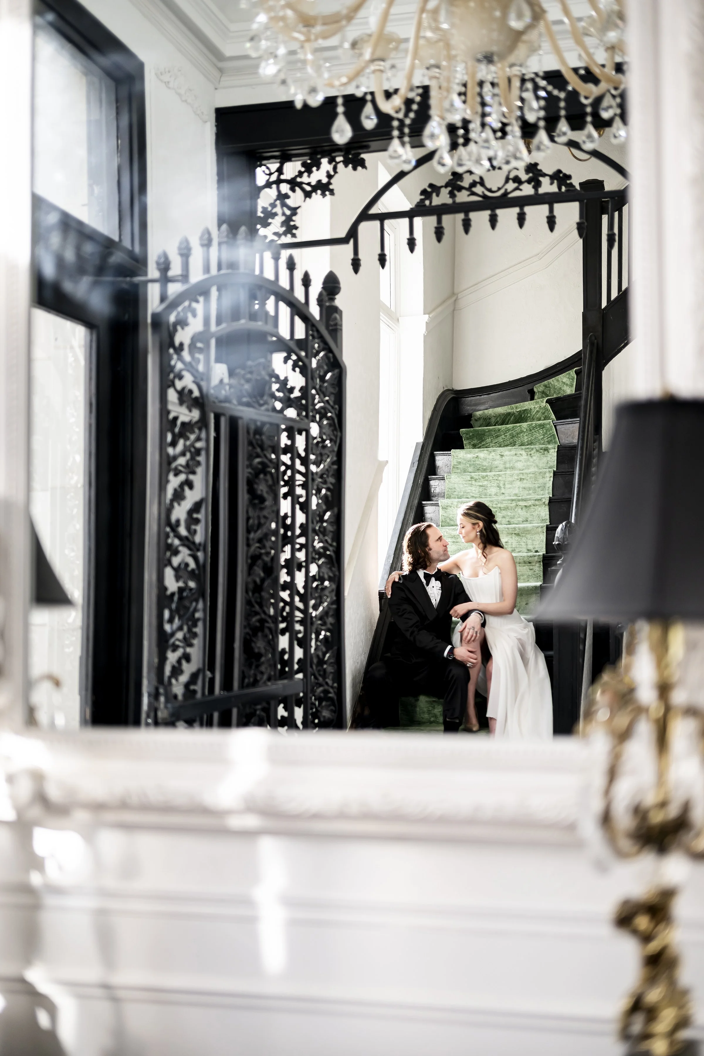 A bride and groom sitting on a staircase, looking at each other, inside a luxurious interior with ornate black iron gates, a chandelier, and large windows, their reflection seen in a nearby mirror.
