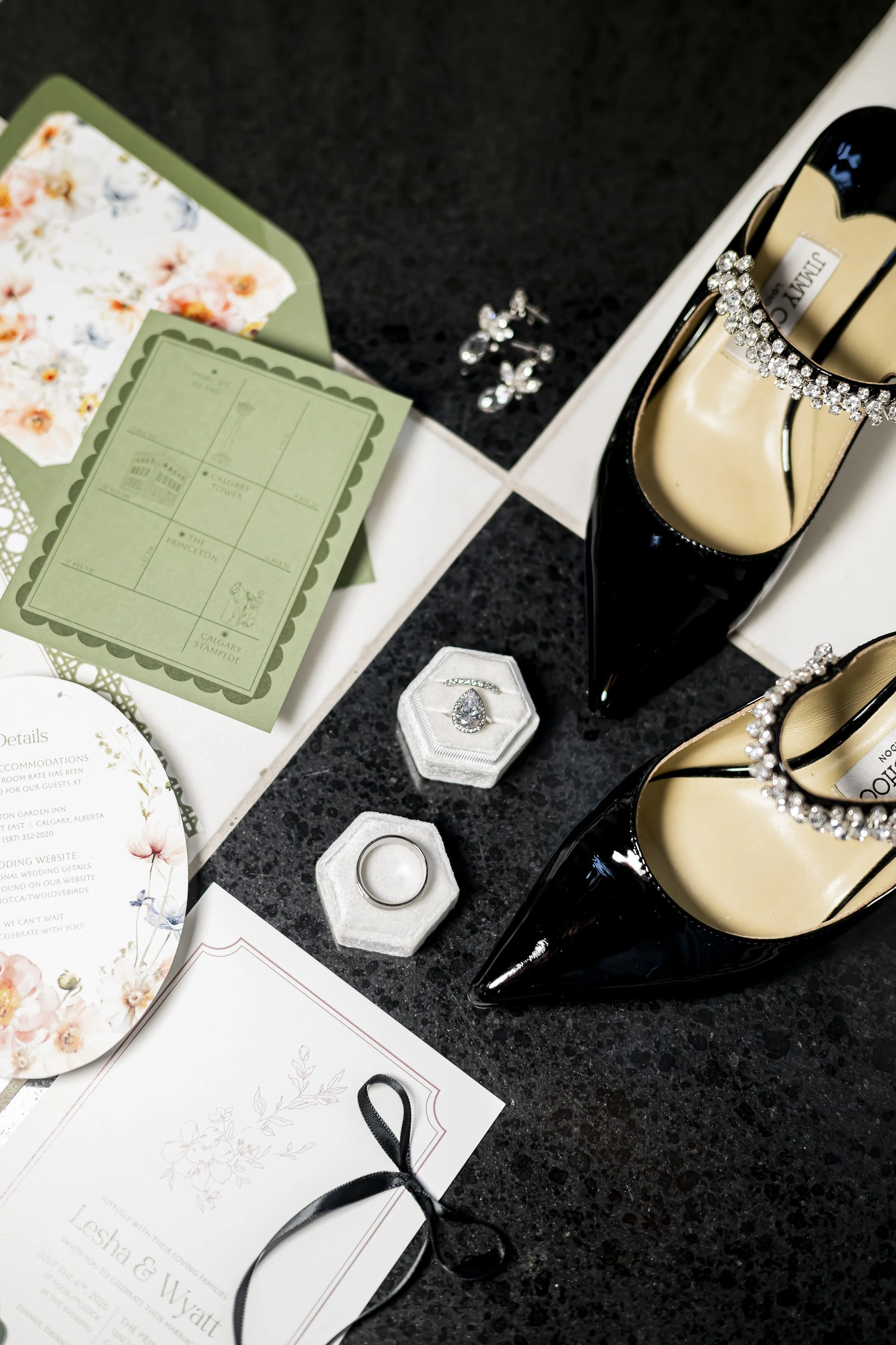 Black high-heeled shoes with rhinestone ankle straps, wedding rings in a jewelry box, and wedding invitations and accessories on a black and white tiled floor.