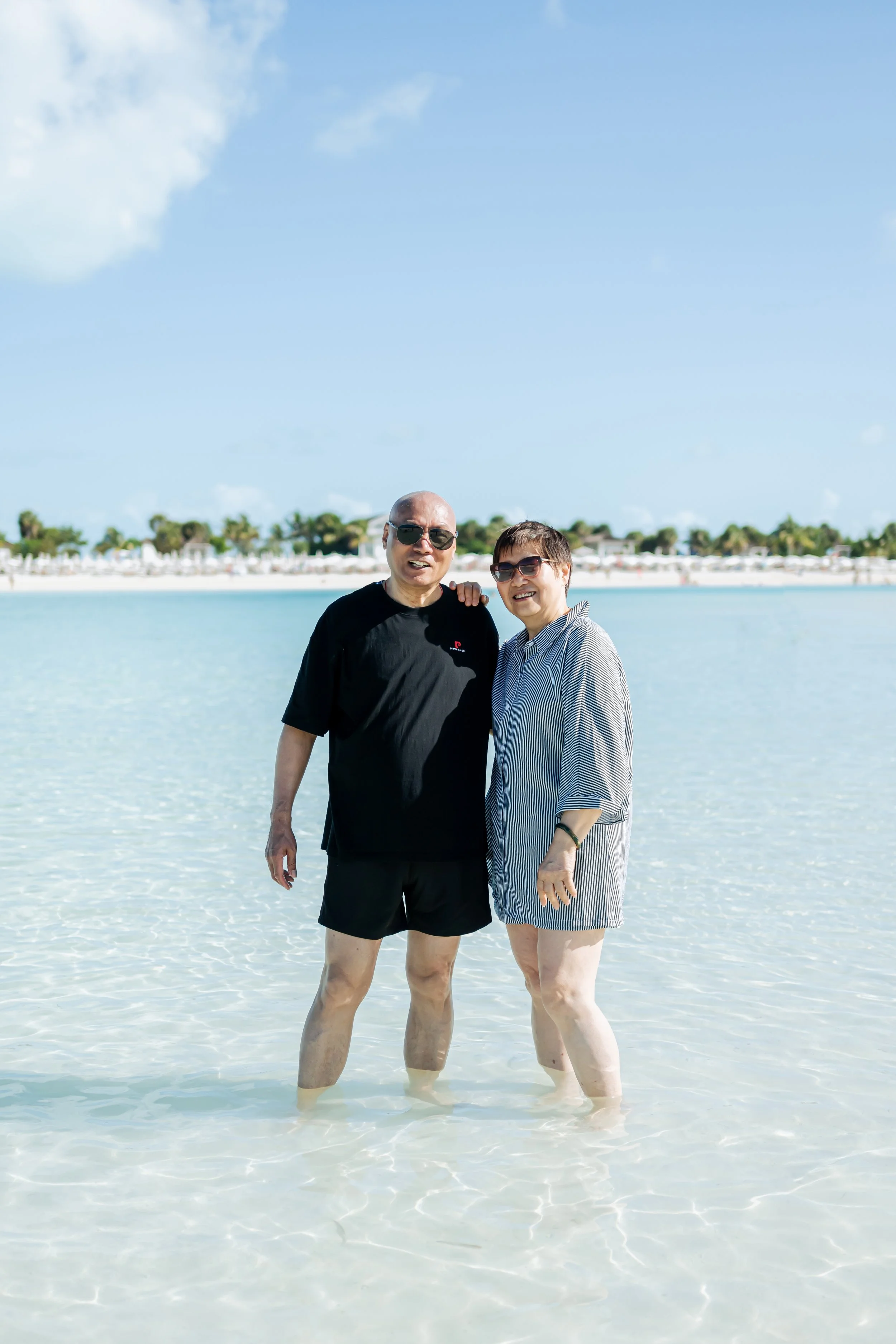 A man and a woman standing barefoot in shallow ocean water, smiling at the camera, with a beach and clear blue sky in the background.