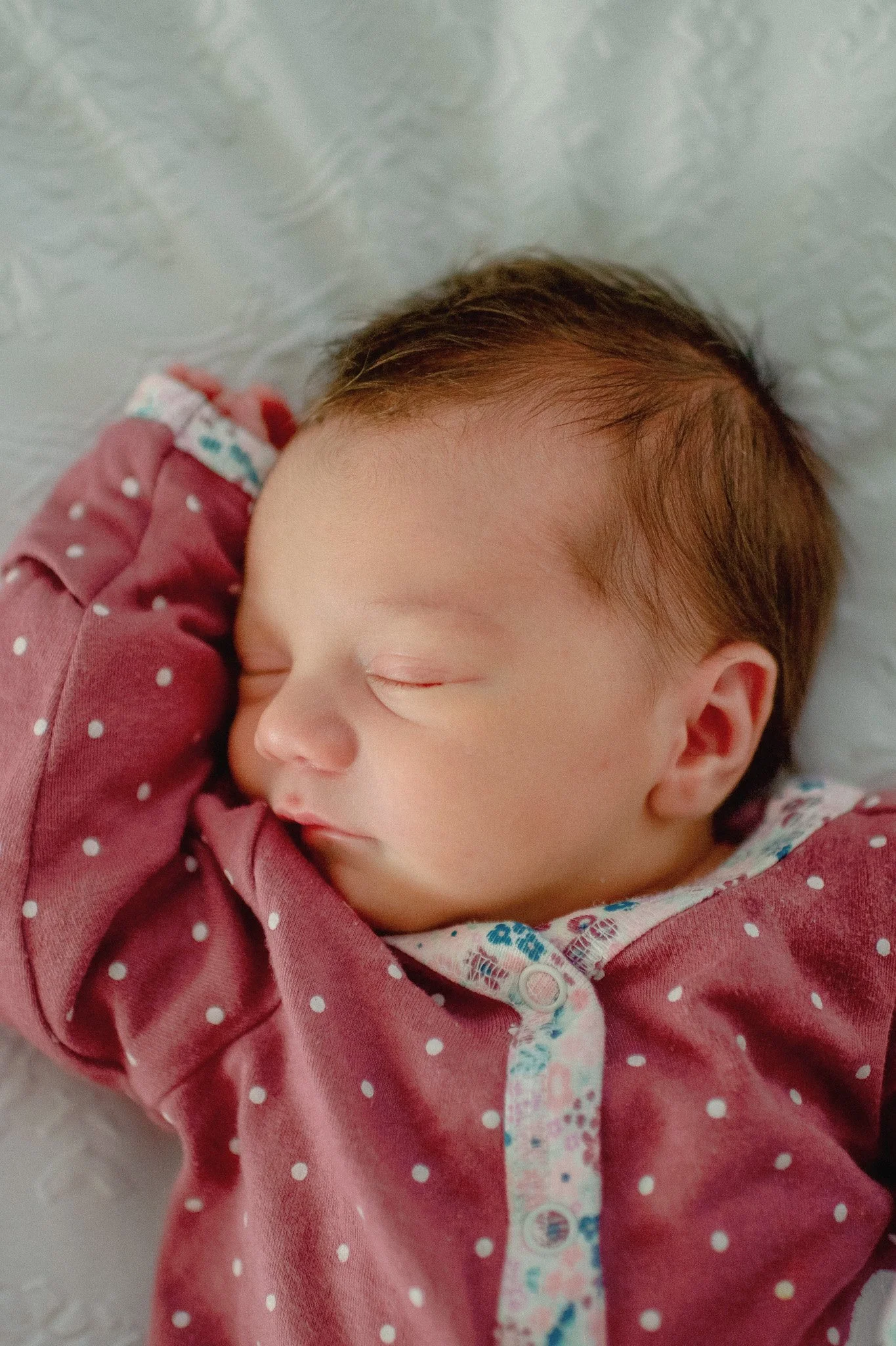 Close-up of a sleeping newborn baby with brown hair, wearing a maroon polka-dot shirt, lying on a soft surface.
