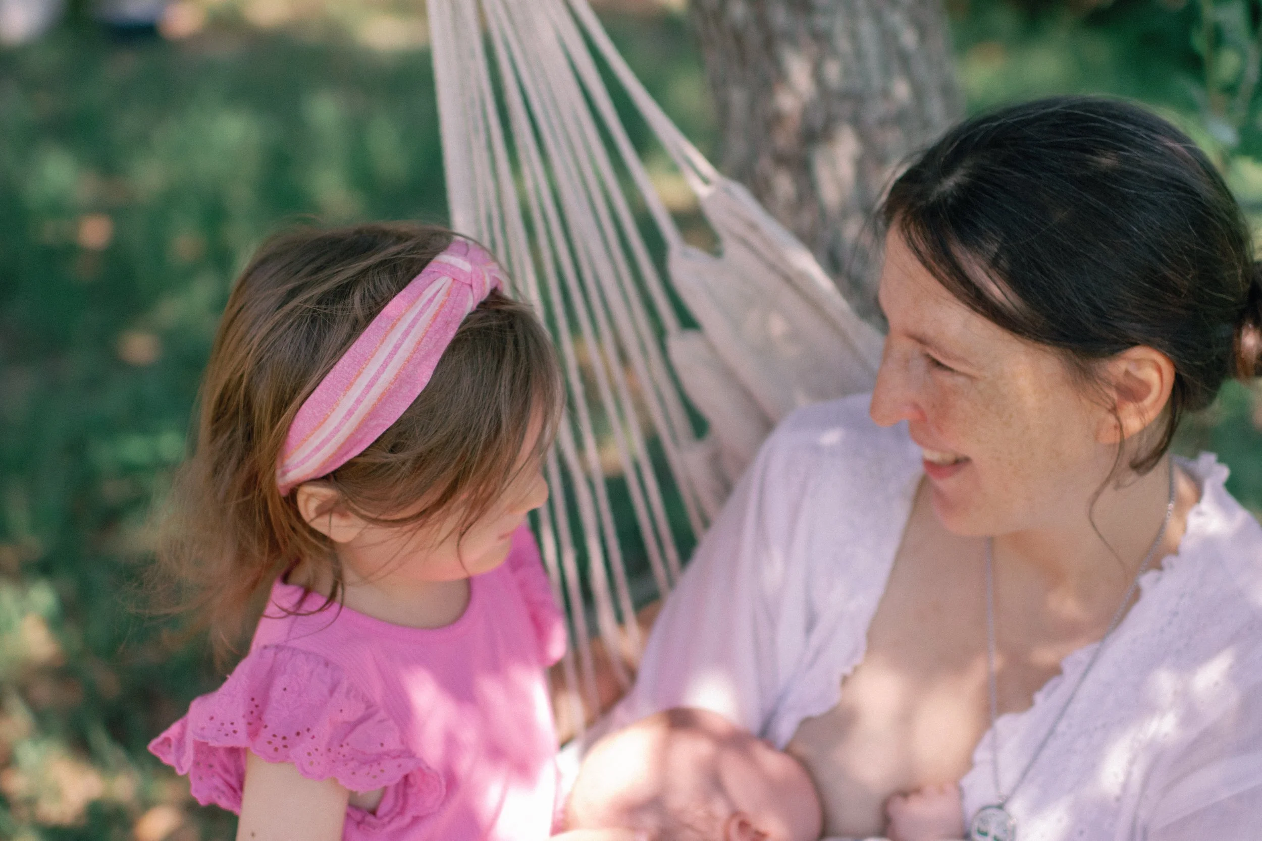 A woman and a young girl smiling at each other outdoors in a park, with a hammock tied between trees in the background.