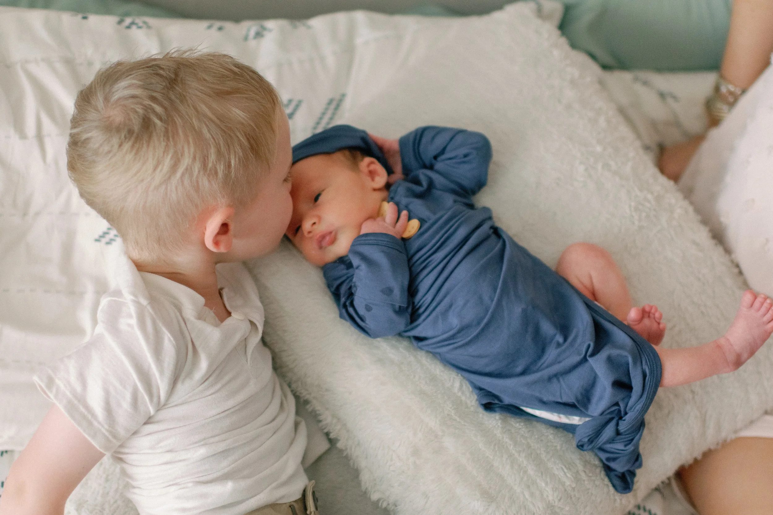 A young boy and a newborn baby lying on a bed, the boy leaning in to kiss the baby on the forehead.