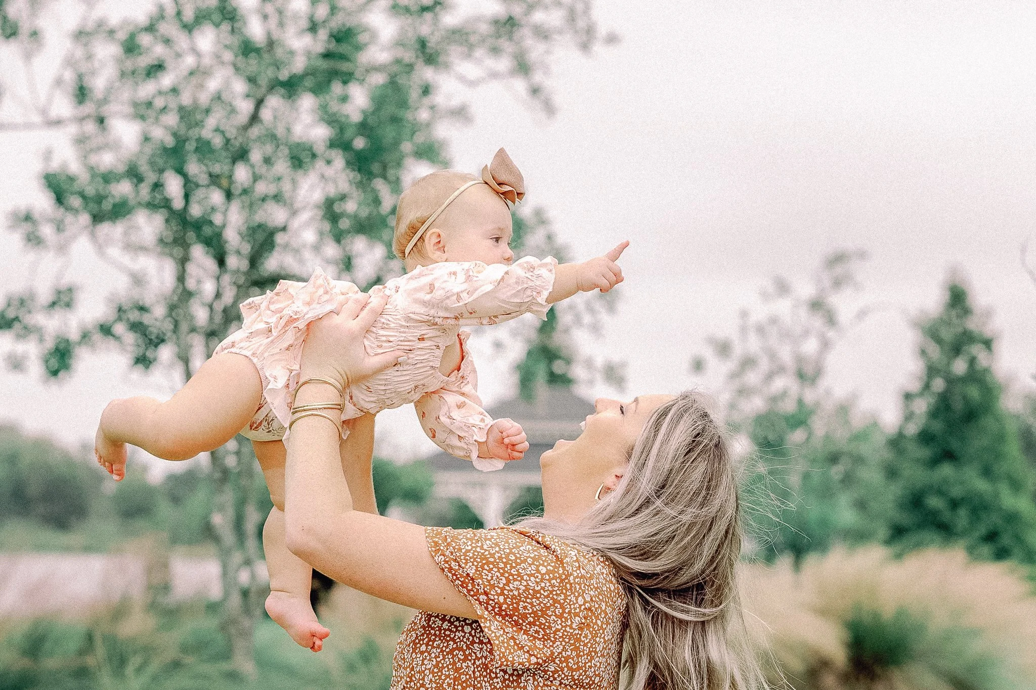 A woman lifts a young girl into the air outdoors, with trees in the background. The girl is pointing and the woman is looking up at her, smiling.