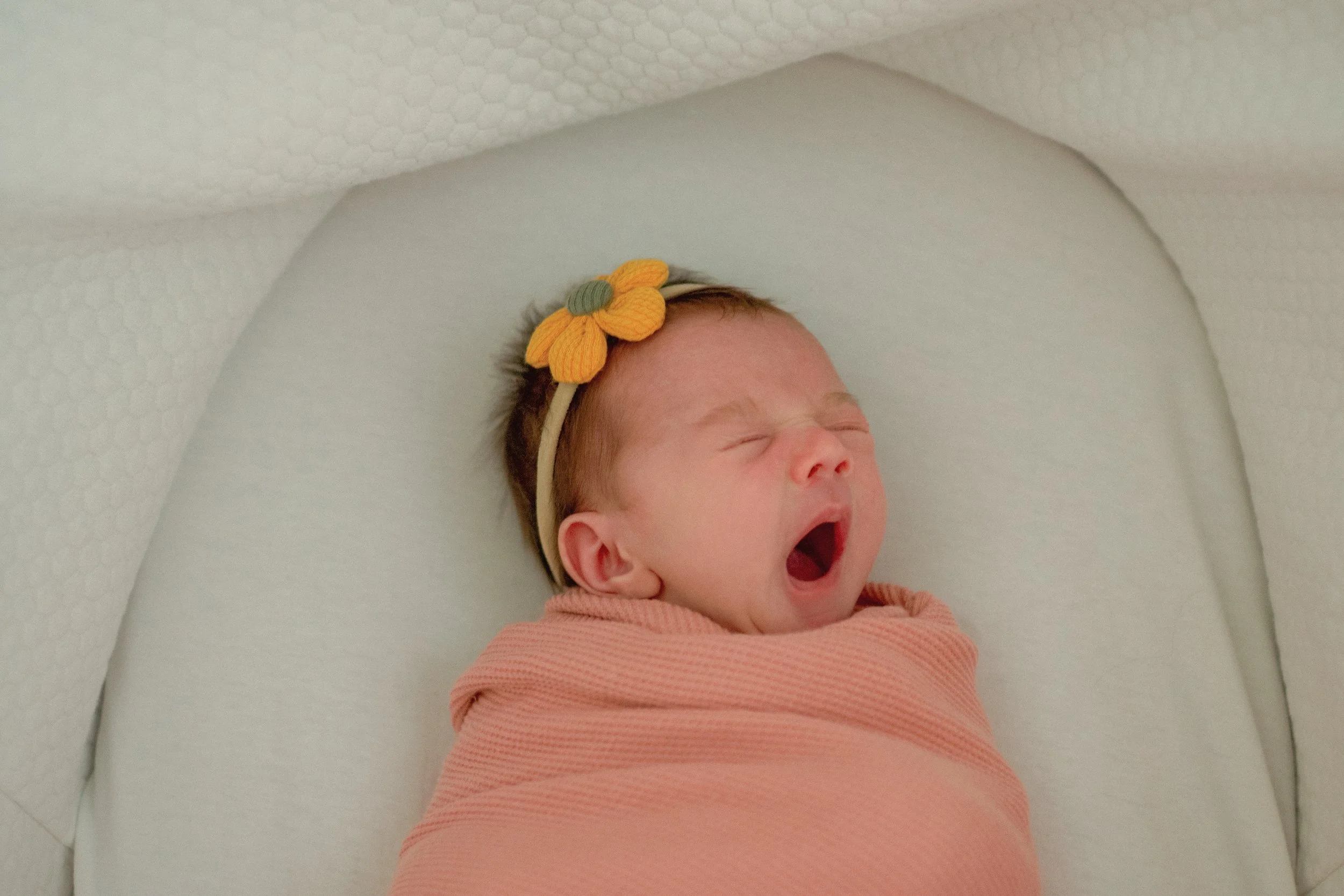 A newborn baby yawning, wrapped in a peach blanket, lying in a white crib with a knitted floral headband featuring a yellow flower attached to it.