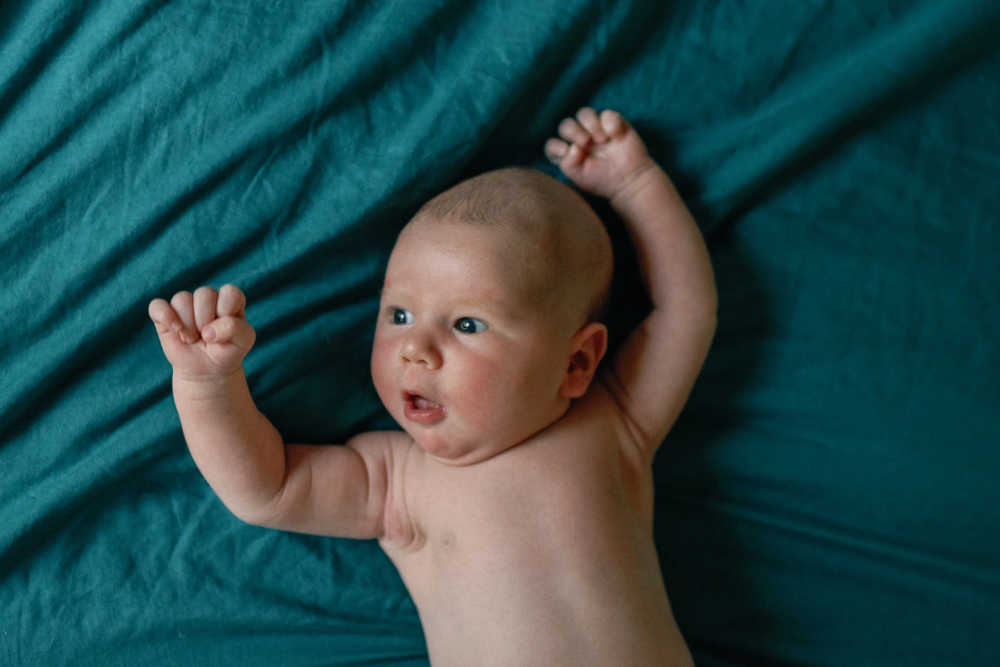 A newborn baby lying on a teal sheet with arms raised and mouth slightly open, gazing to the side