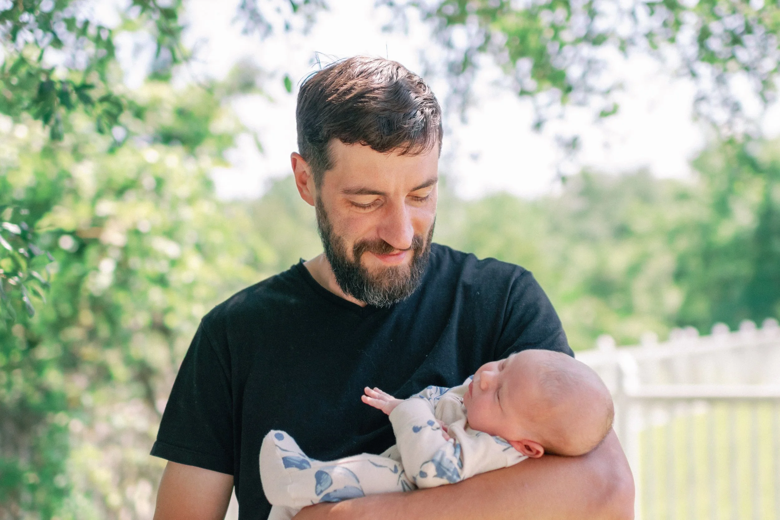 A man with a beard and short brown hair holding a baby wrapped in a blanket outside, with trees and a fence in the background.