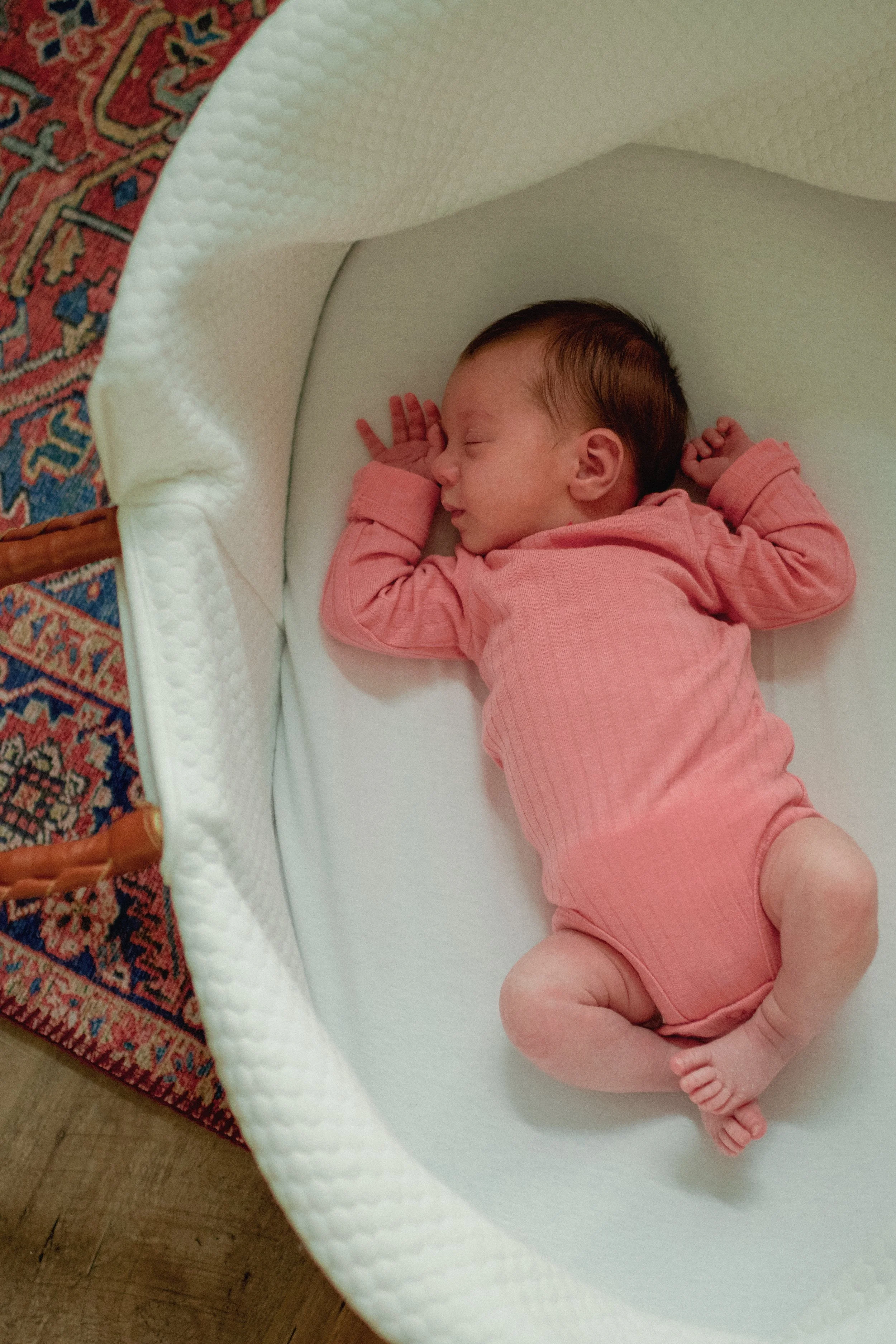 A sleeping baby with a pink outfit sleeping in a white bassinet with a textured pattern. The bassinet is placed on a patterned rug.