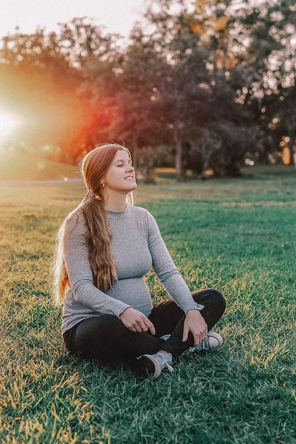 A pregnant woman sitting on grass with eyes closed in a park during sunset.