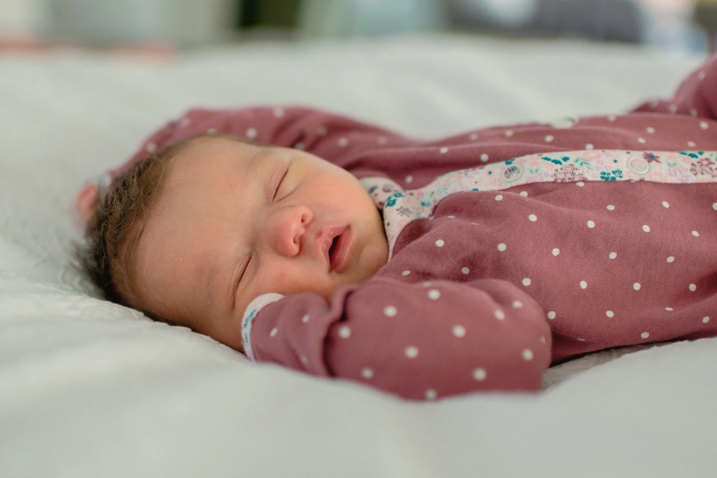 A sleeping baby lying on a white bed, wearing a pink polka dot onesie.