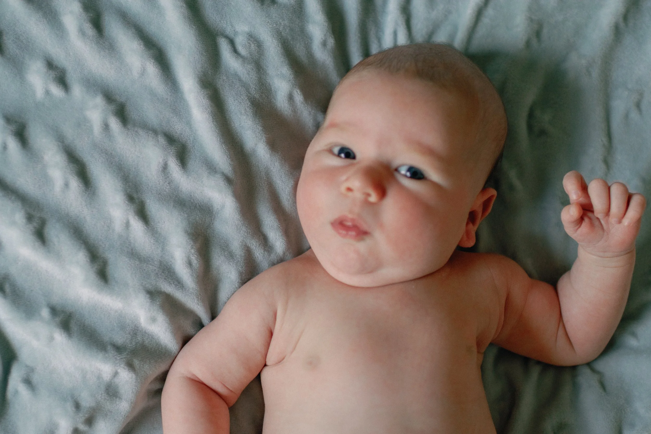 A baby lying on a soft, grey blanket, looking up at the camera, with one arm raised and a curious expression on their face.