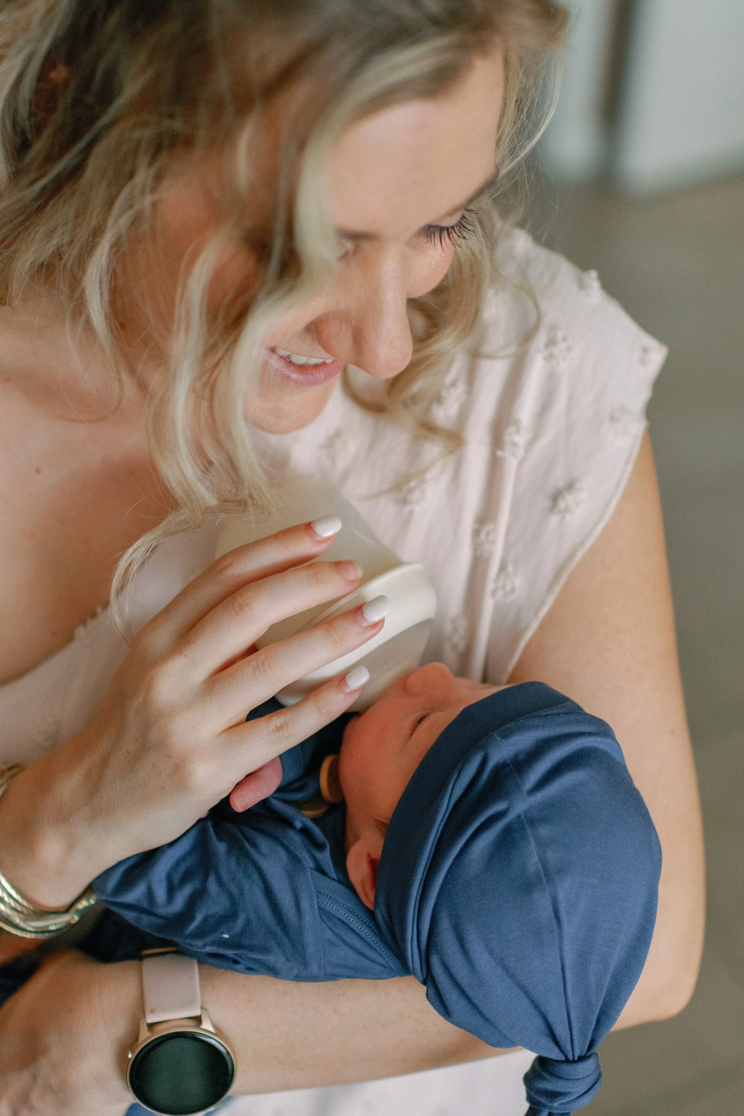 A woman with blonde hair feeding a baby with a bottle, the woman is smiling and wearing a light-colored top, the baby is dressed in a blue hat and clothing.