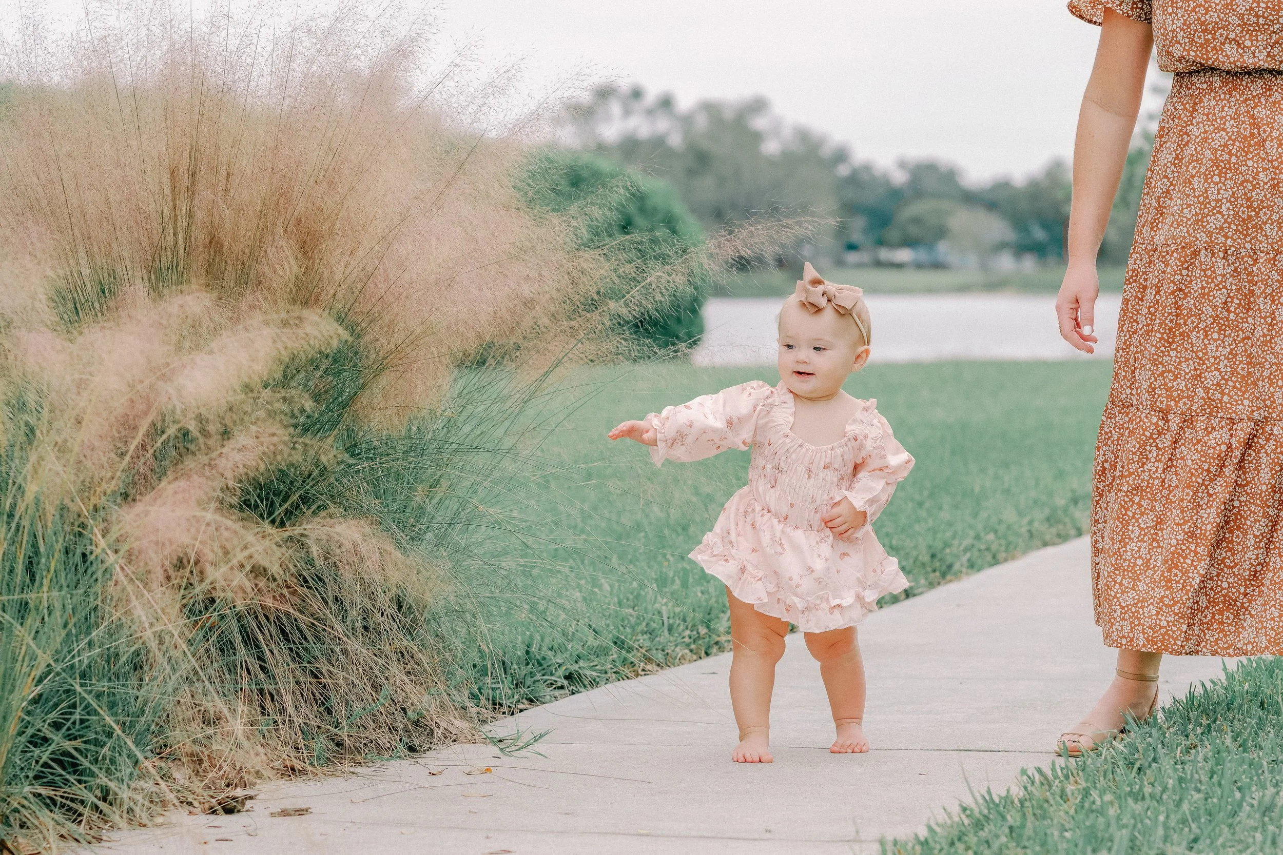 A young girl in a pink floral dress with a large bow in her hair is walking barefoot on a sidewalk next to a woman, who is wearing a long orange floral dress and sandals. They are outdoors near a body of water with tall ornamental grass on the side of the sidewalk.