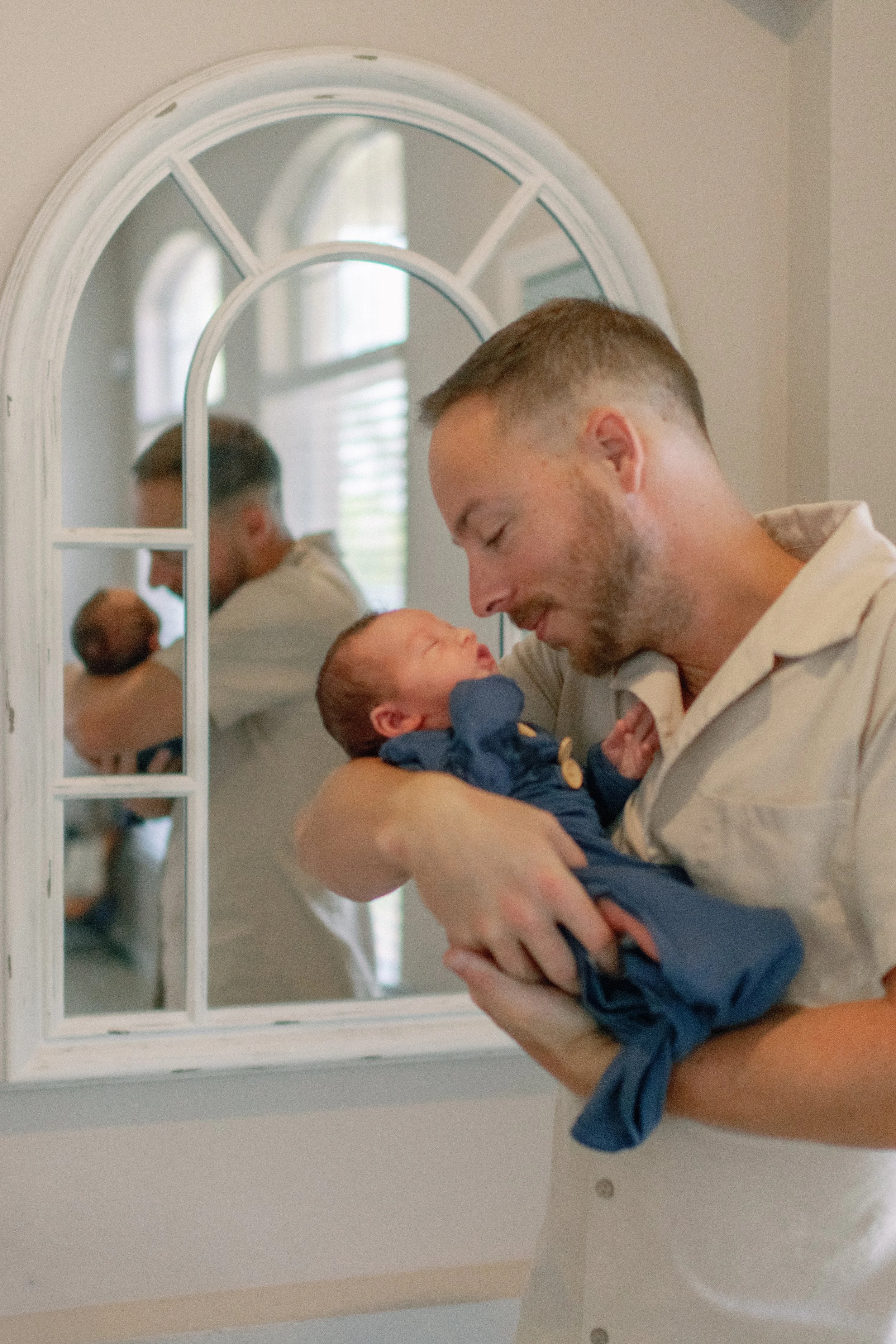 A man holding a newborn baby in front of a mirror, both peacefully smiling.
