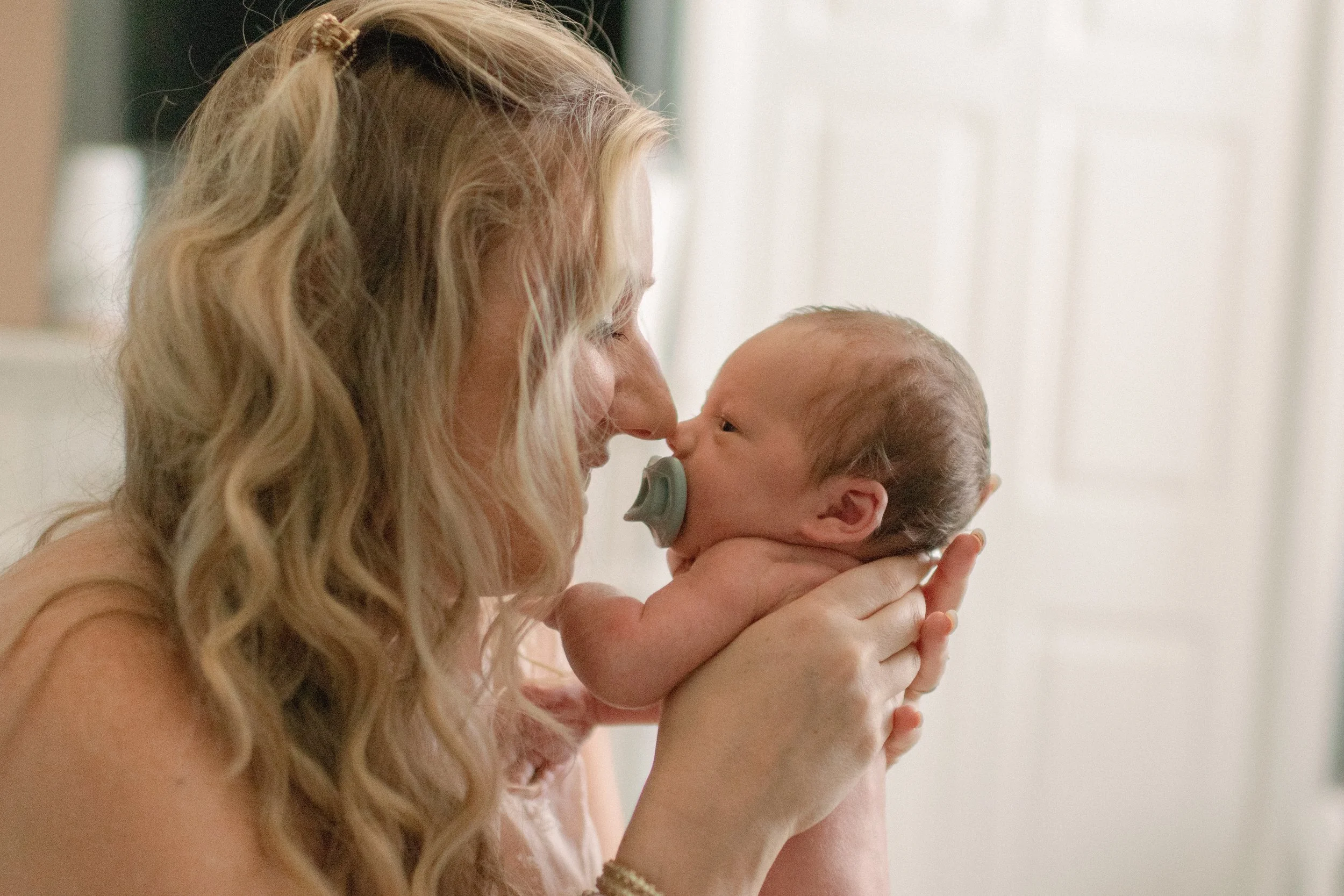 A woman with blonde wavy hair holding a newborn baby with a pacifier in its mouth, both touching noses in a tender moment indoors.
